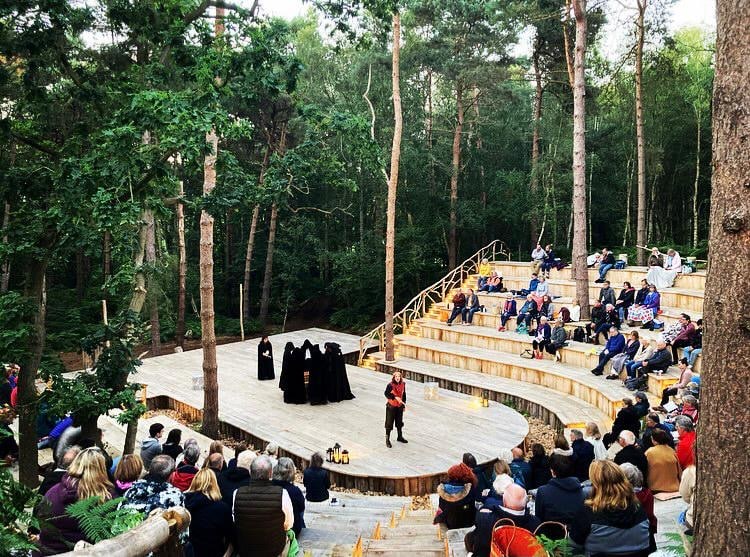 A captivating outdoor theatre performance at Thorington Theatre, an open-air amphitheater nestled in a woodland setting. The audience sits on tiered wooden seating, attentively watching a scene unfold on the circular wooden stage. Several actors dressed in long black cloaks stand in a solemn formation, while another performer in a red and black costume addresses them. Lanterns placed around the stage add to the atmospheric setting, surrounded by tall trees and lush greenery.