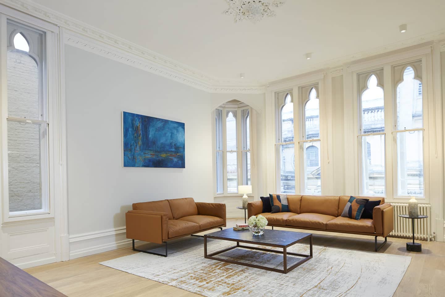 High Street Apartment living room, featuring two brown sofas, a coffee table, a radiator, a blue painting, and several large windows