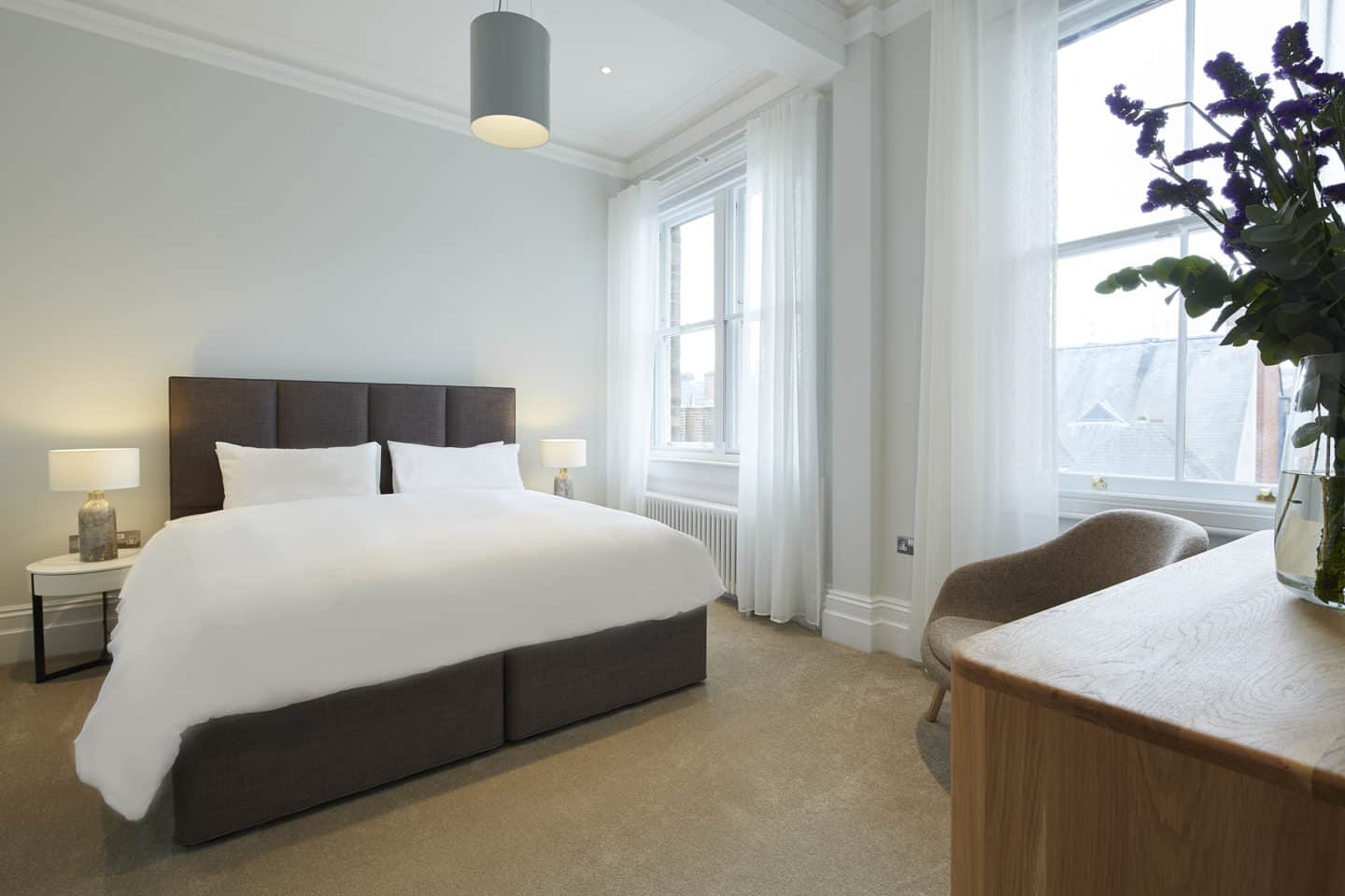 Photograph of a High Street Apartment, showing a bed with white sheets and pillows, two lamps on bedside tables, a table, an armchair, a radiator, and two large windows