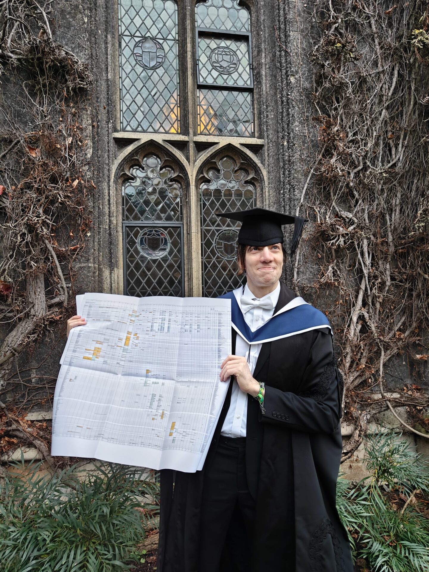 Photo of René Lavanchy wearing graduation gown and mortar board holding a newspaper facing outwards. He's standing in front of a window in Front Quad.