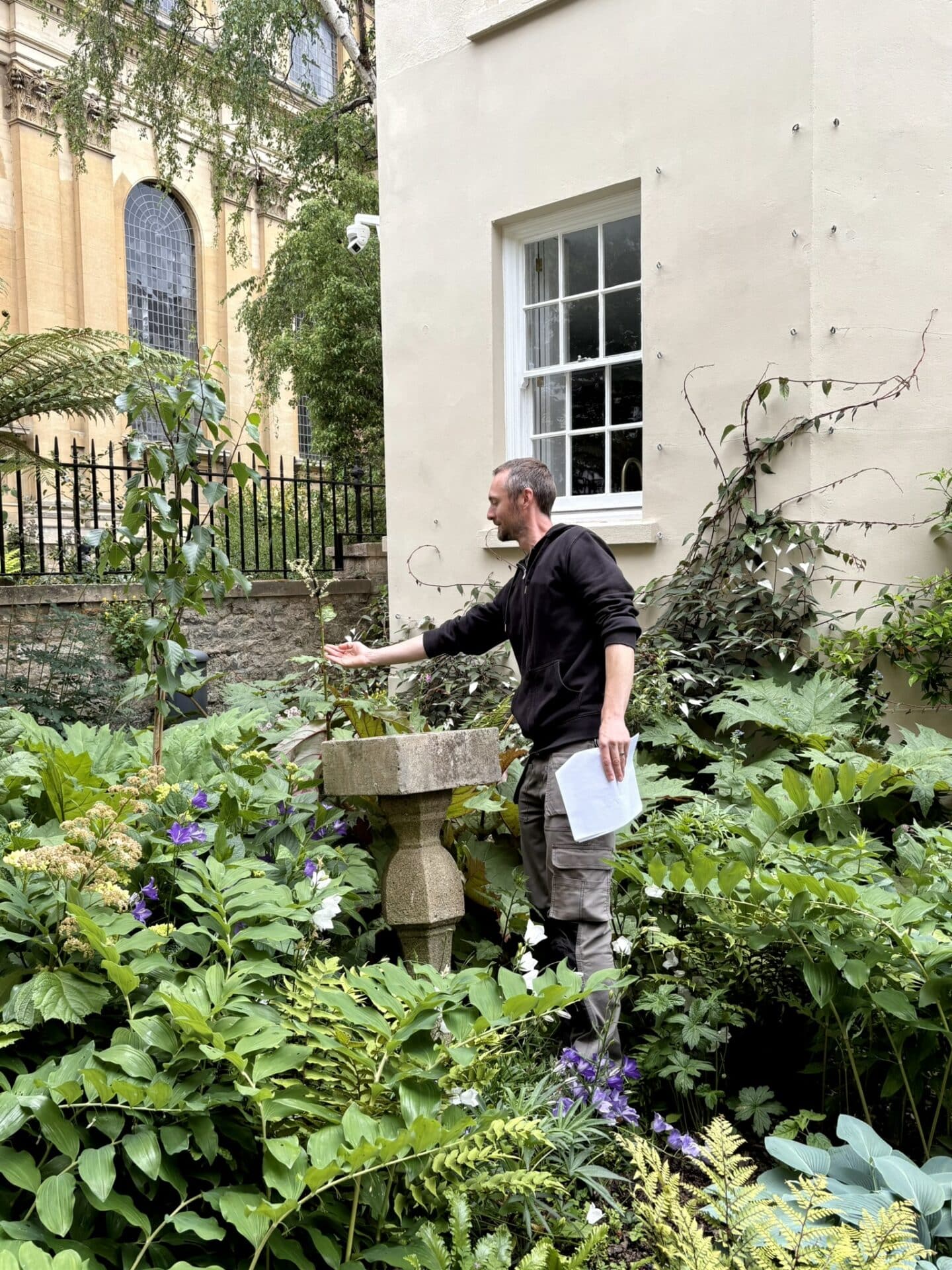 Mike Hawkins gestures towards planting in Lincoln College’s Fellows’ Garden, surrounded by lush greenery and architectural stonework. The main library is visible in the background.