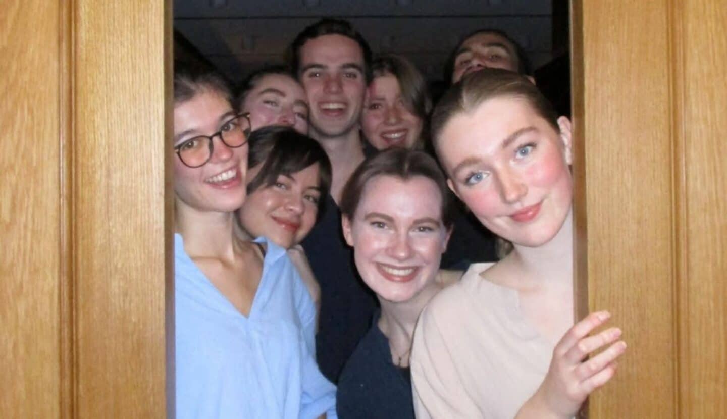 A group of smiling cast members crowd together in a doorway