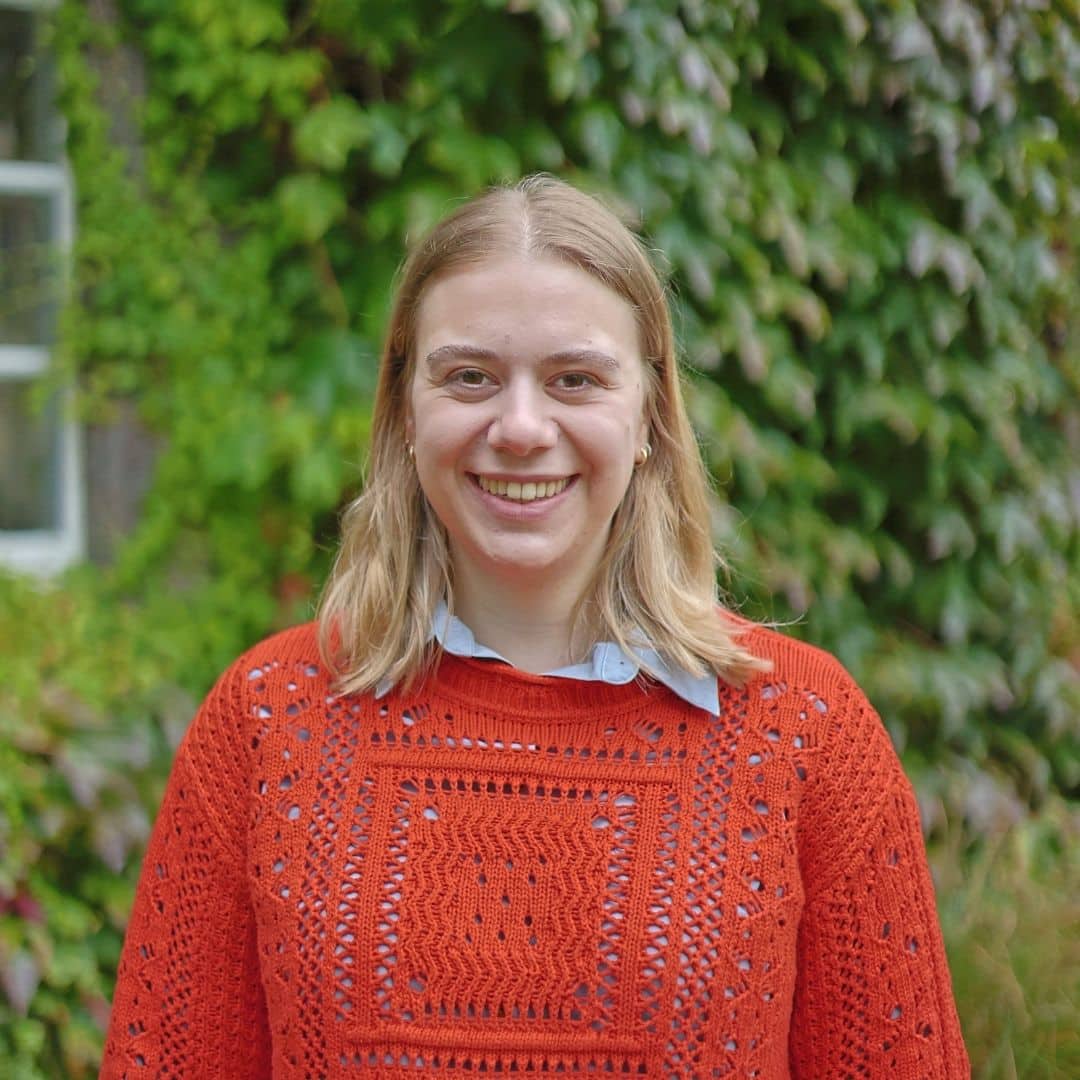Photo of Juliet Tye in an orange jumper standing in front of ivy