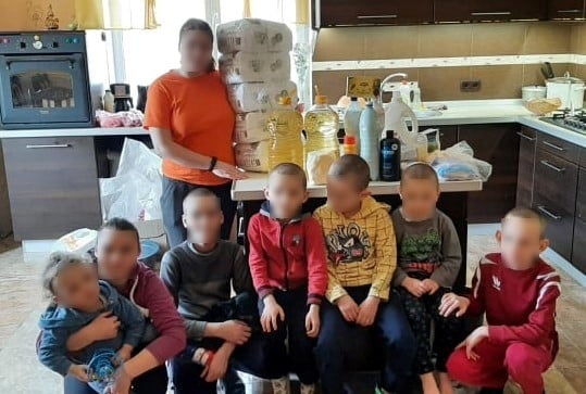 A family of 8 in a kitchen, with a pile of groceries on the table. Faces are blurred out.