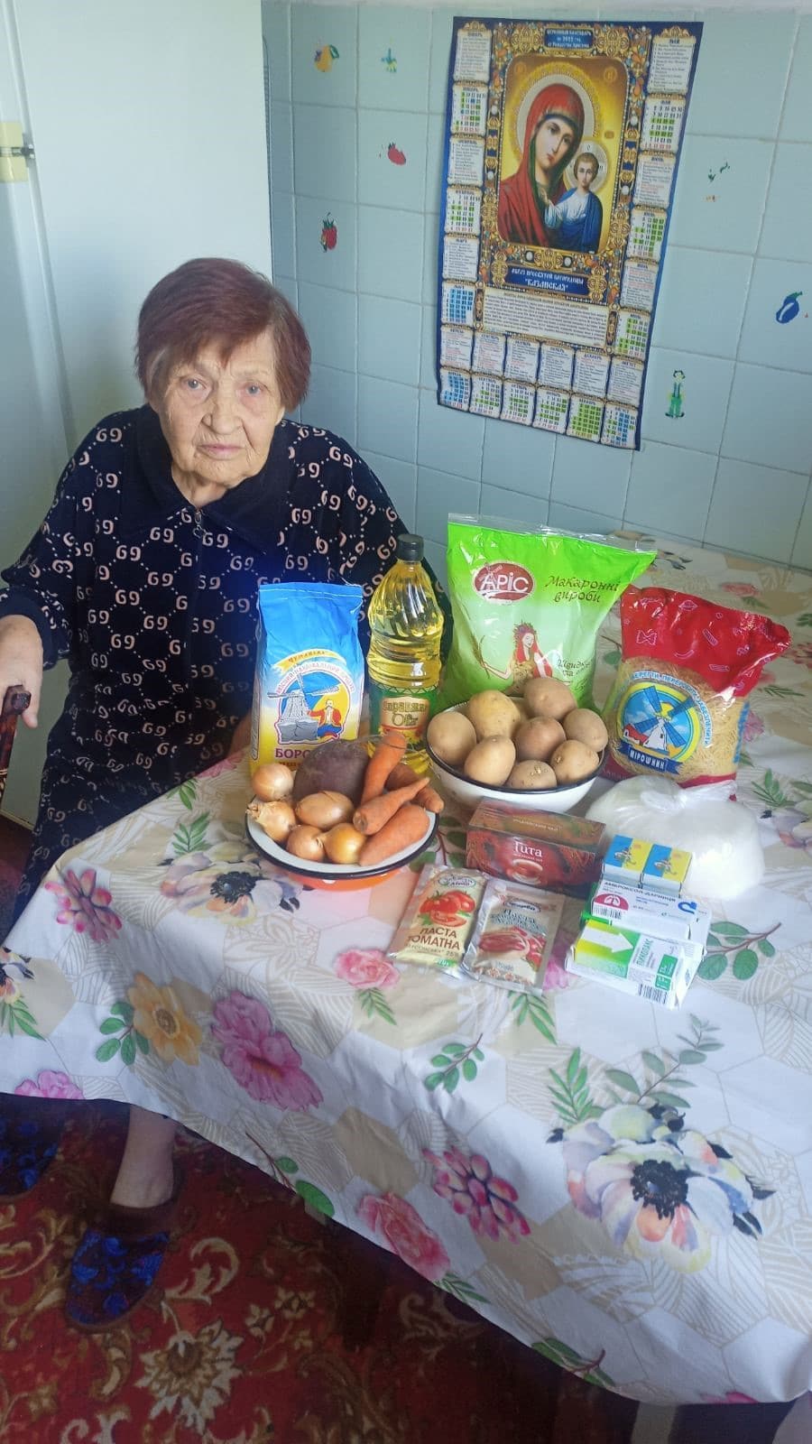 An elderly woman sitting in a kitchen, with a pile of groceries on the table in front of her.