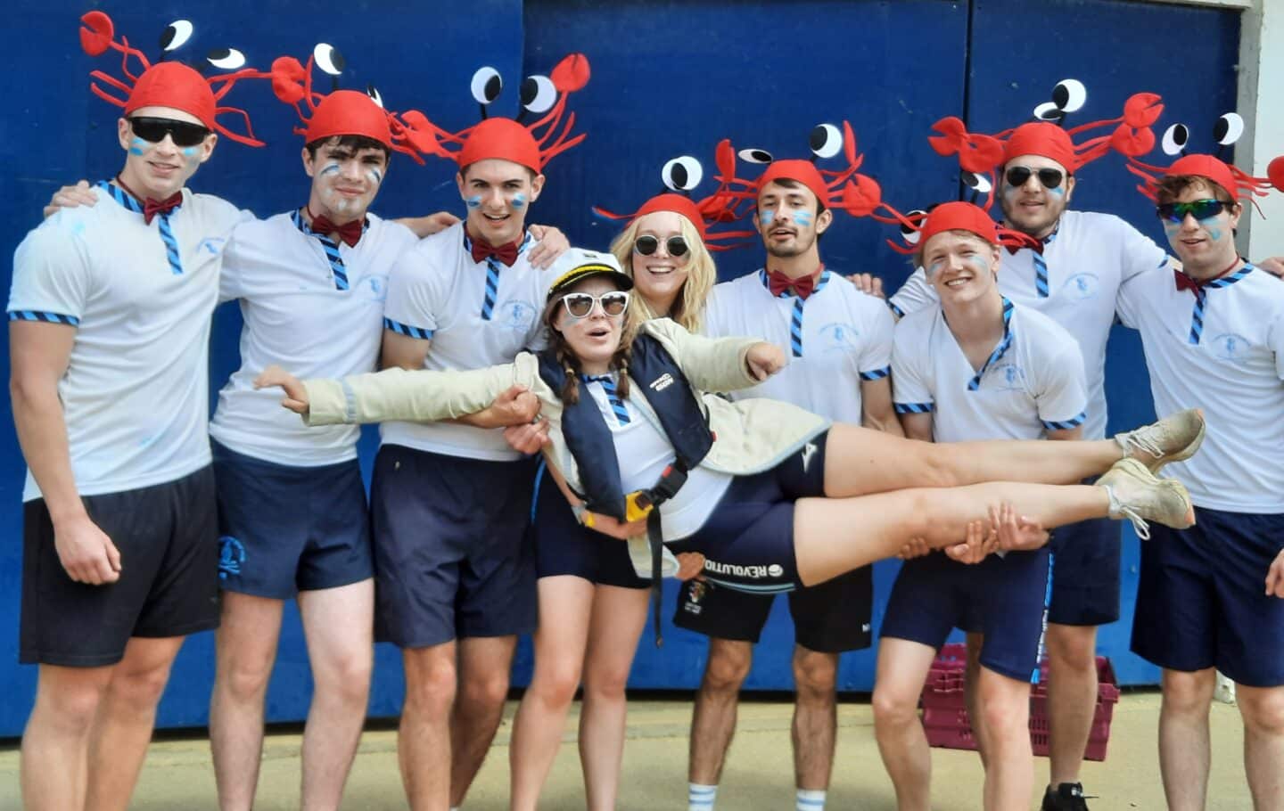 M2 crew posing in a line holding up crew member. All are in kit and wearing fun red lobster hats