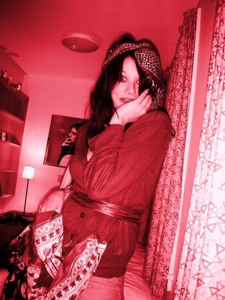 A stylized red-tinted photograph of Beckis Cooper in a Lincoln College, Oxford dormitory room. She is dressed in a bohemian-inspired outfit, wearing a leopard-print headscarf and a layered ensemble with patterned scarves tied around her waist. She poses playfully with one hand on her face. The background features shelves, a desk lamp, and a poster of a woman smoking, with patterned curtains framing the room.