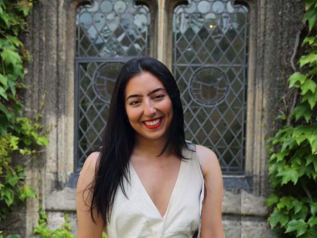 Maria Murad smiling, wearing a formal white dress and standing in front of a stained glass window