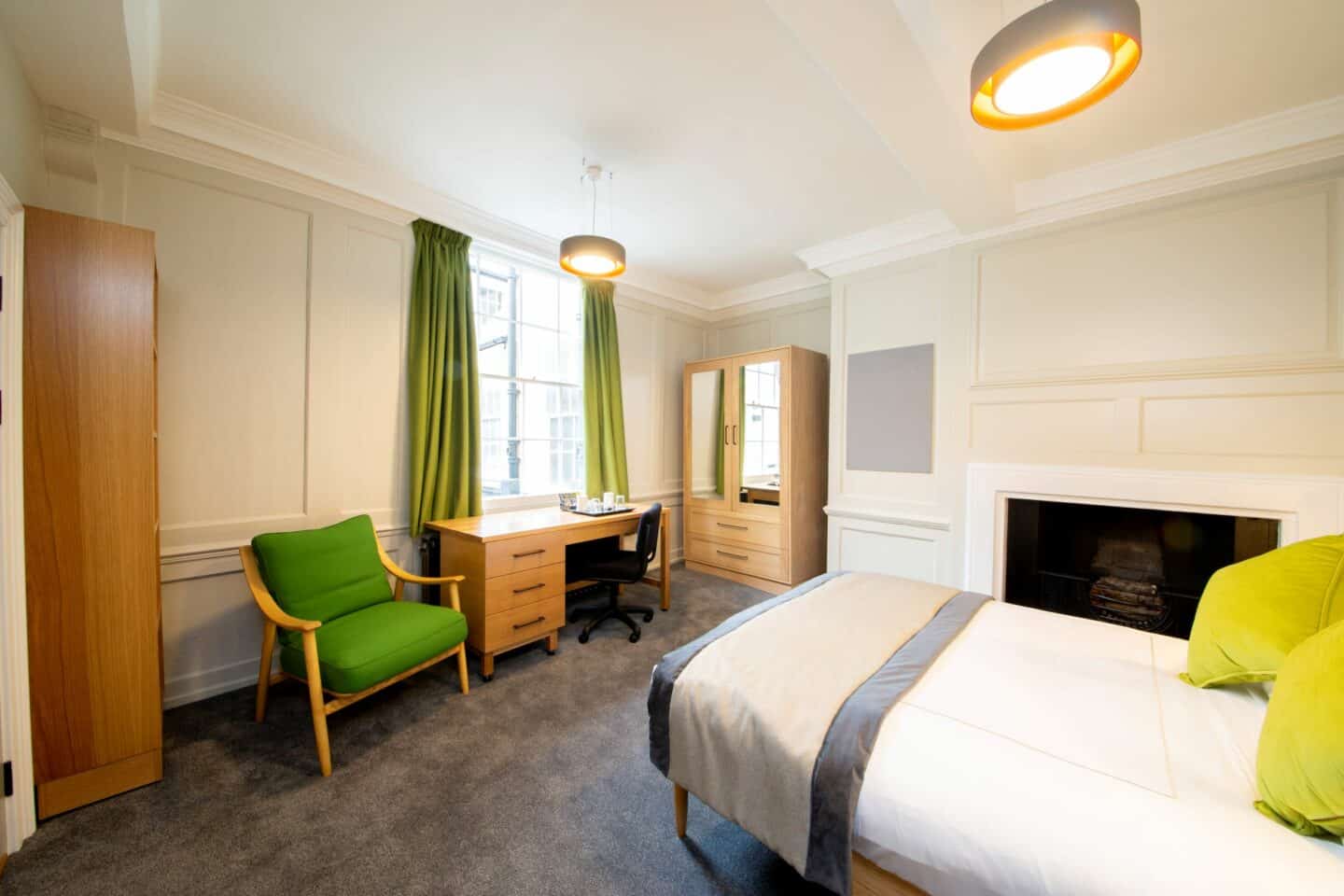 Bedroom with a double bed (with sheets, pillows, and green cushions), an armchair, a wooden desk with chair, drawers, and tea-set, a radiator, a set of shelves, a wardrobe with drawers and mirrors, a corkboard, a fireplace, and a window with curtains