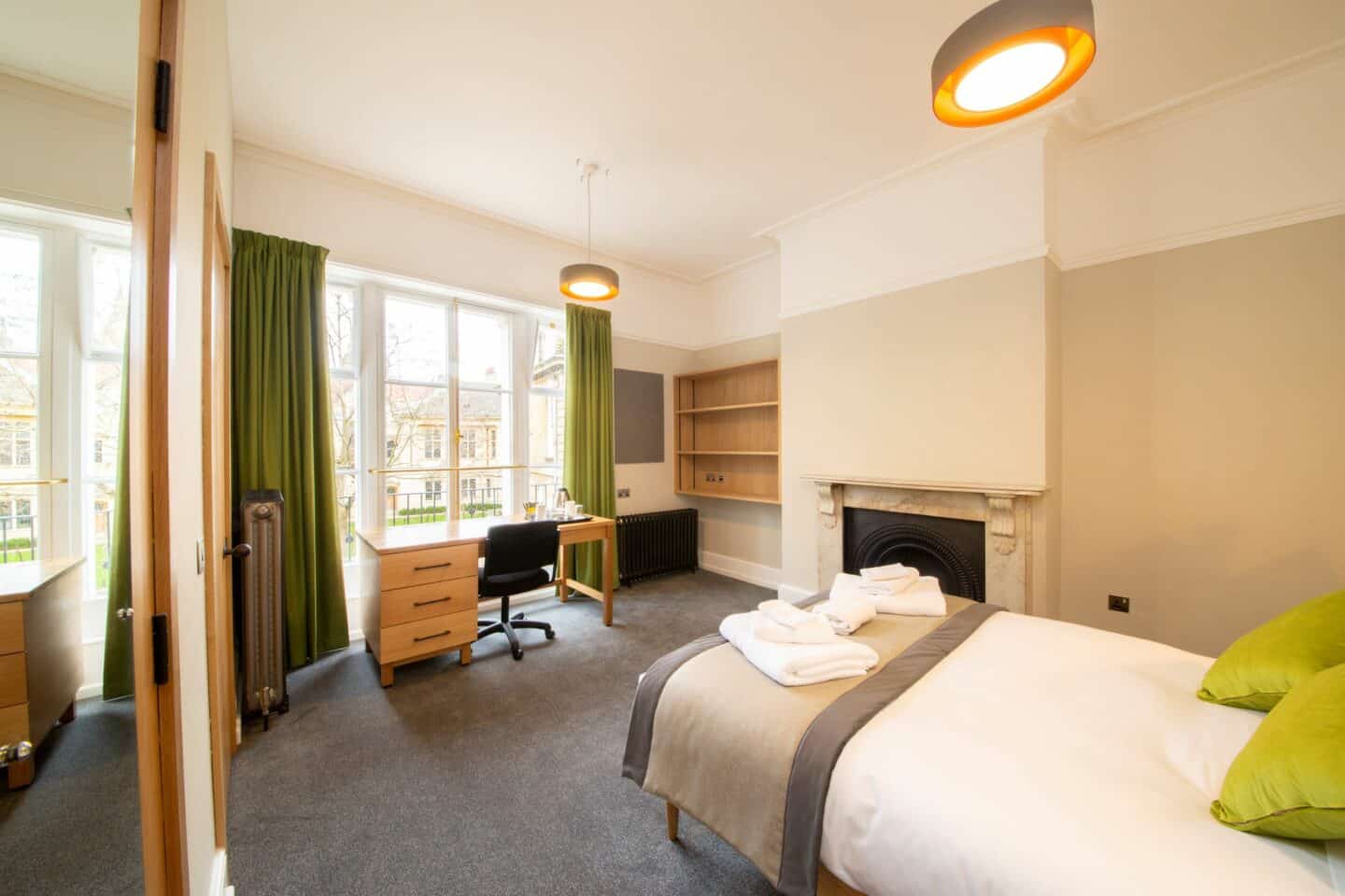 F78 - bedroom with a single bed (with sheets, cushions, and towels), a fireplace, shelves, a corkboard, a radiator, a wooden desk with drawers, chair, kettle and tea-set, large windows with green curtains, a mirror and a door