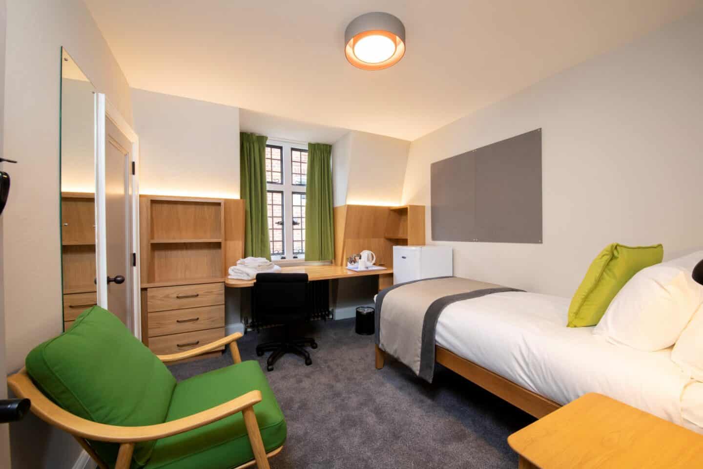 Bedroom with a single bed (with pillows, a cushion and sheets), corkboards, a bedside table, an armchair, a chest of drawers and shelves, a mirror, a wooden desk with chair, shelves, towels, kettle and tea-set, a bin, a mini-fridge, and windows with curtains