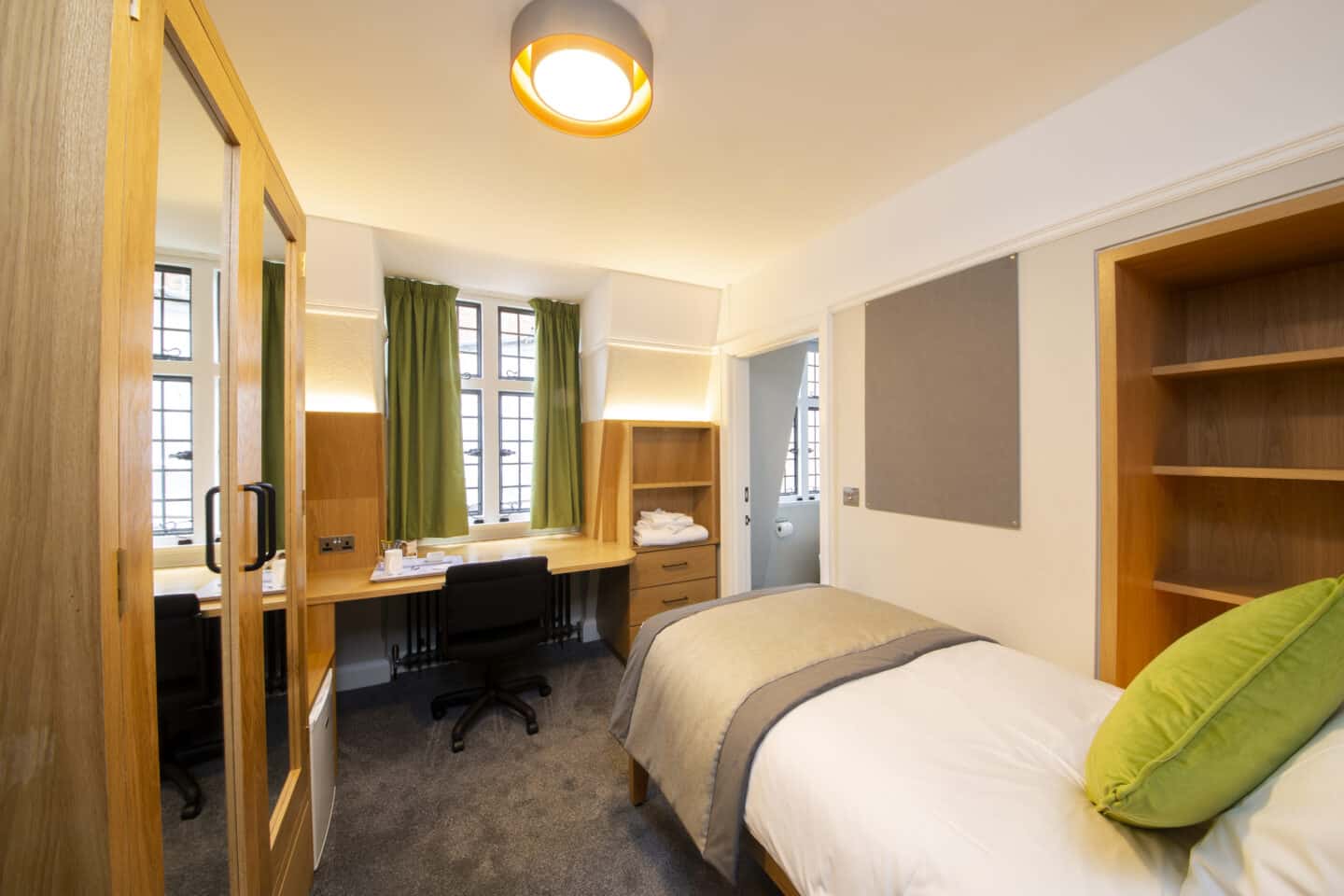 Bedroom with a single bed (with pillow, cushion and sheets), a large wardrobe with mirrors, a set of shelves, a wooden desk with chair, drawers, shelves, and tea-set, towels, windows with curtains, a corkboard, a radiator and an en-suite bathroom