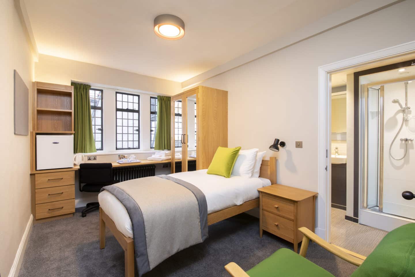 Bedroom with a single bed (with sheets, pillows and a cushion), a small chest of drawers, a wardrobe with mirrors, a desk with chair, shelves, drawers, kettle, tea-set and towels, large windows with green curtains, a corkboard, a mini-fridge, an armchair, and an en-suite bathroom with shower and sink visible
