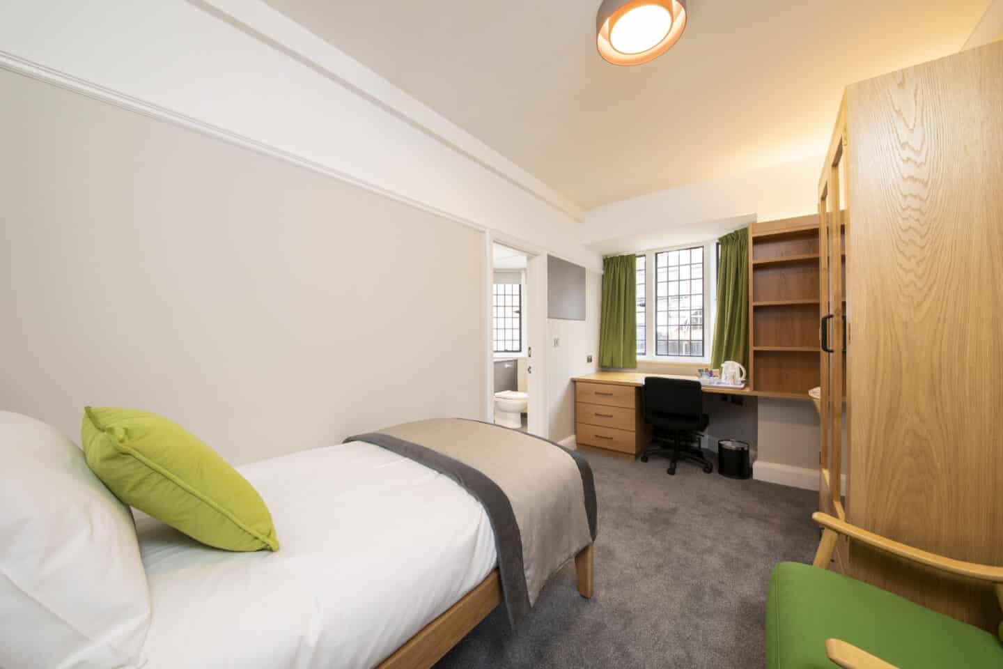 Bedroom with a single bed (with pillow, cushion and sheets), an armchair, a large wardrobe with mirrors, a set of shelves, a wooden desk with drawers, chair, kettle and tea-set, a bin, windows with curtains, a corkboard, and an en-suite bathroom with toilet visible