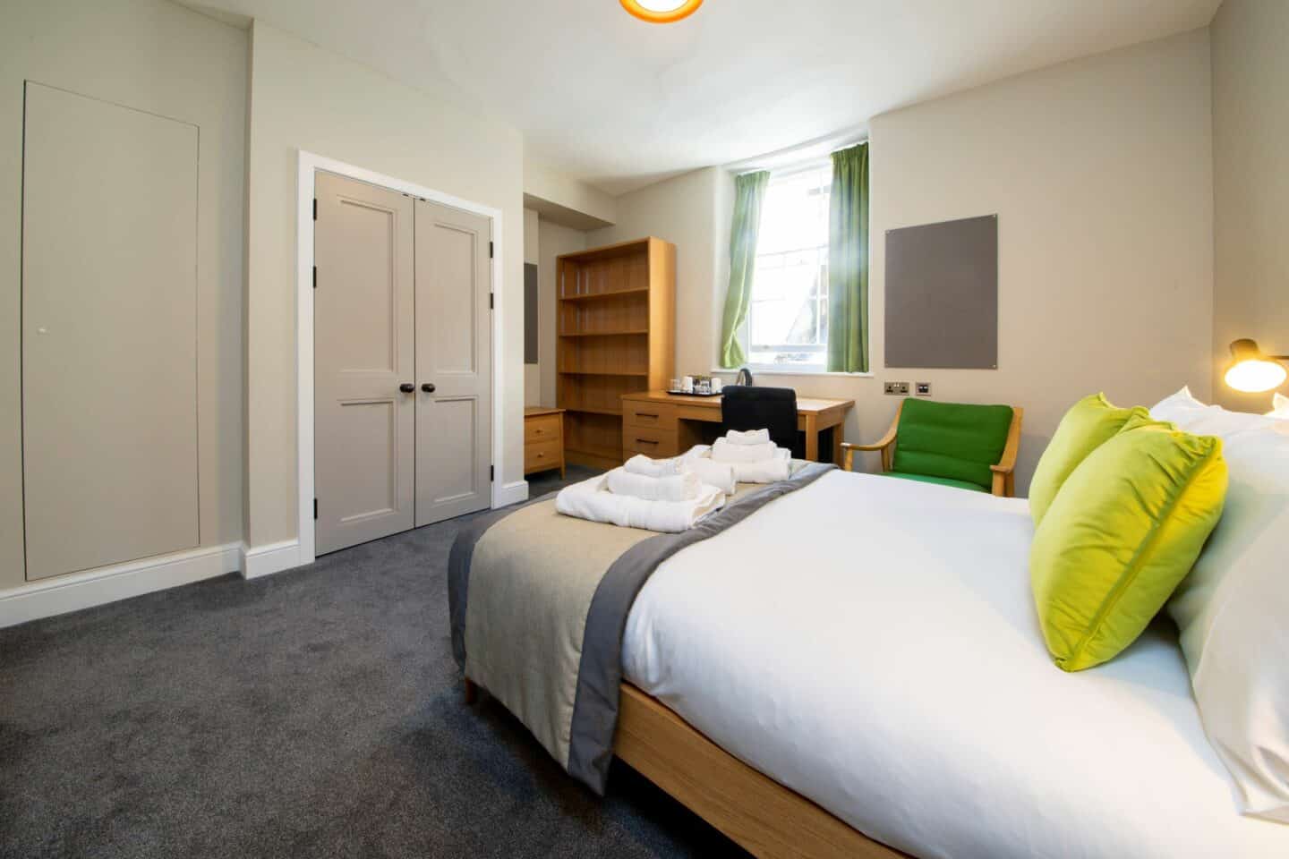 F40 - bedroom with a double bed (with sheets, towels, pillows and cushions), an armchair, a corkboard, a window with curtains, a wooden desk with chair, drawers, a kettle and tea-set, a set of shelves, a small chest of drawers, and a set of double doors