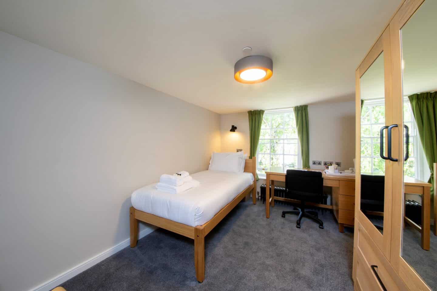 S52 - bedroom with a single bed (with sheets, pillows, towels, and toiletries), a radiator, a wooden desk with chair, drawers, kettle and tea-seat, a large wardrobe with mirrors and drawers, and a large window with curtains