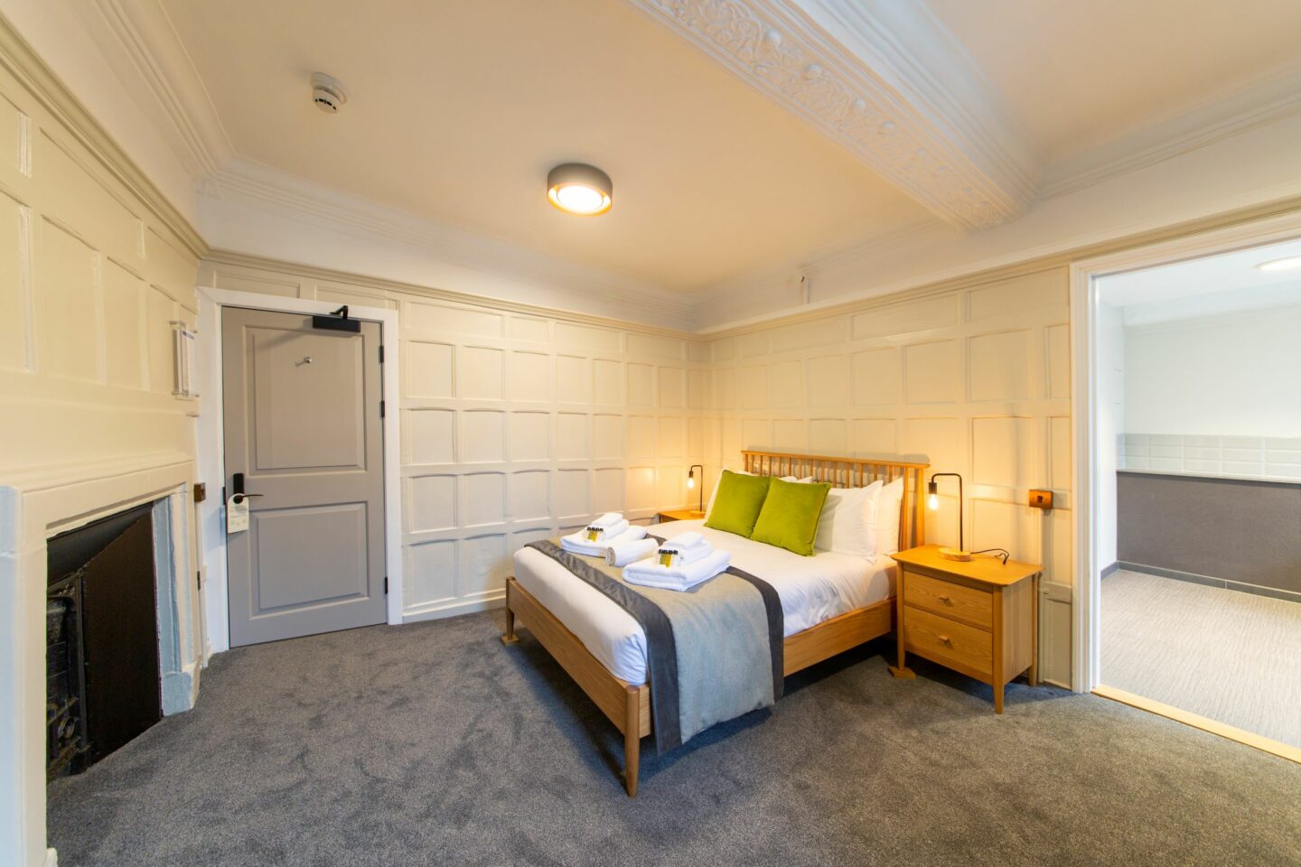 S22 - bedroom with white panelled walls, a double bed (with pillows, cushions, sheets, towels and toiletries), a fireplace, two bedside tables with drawers and lamps, and a large doorway opening up into a different room