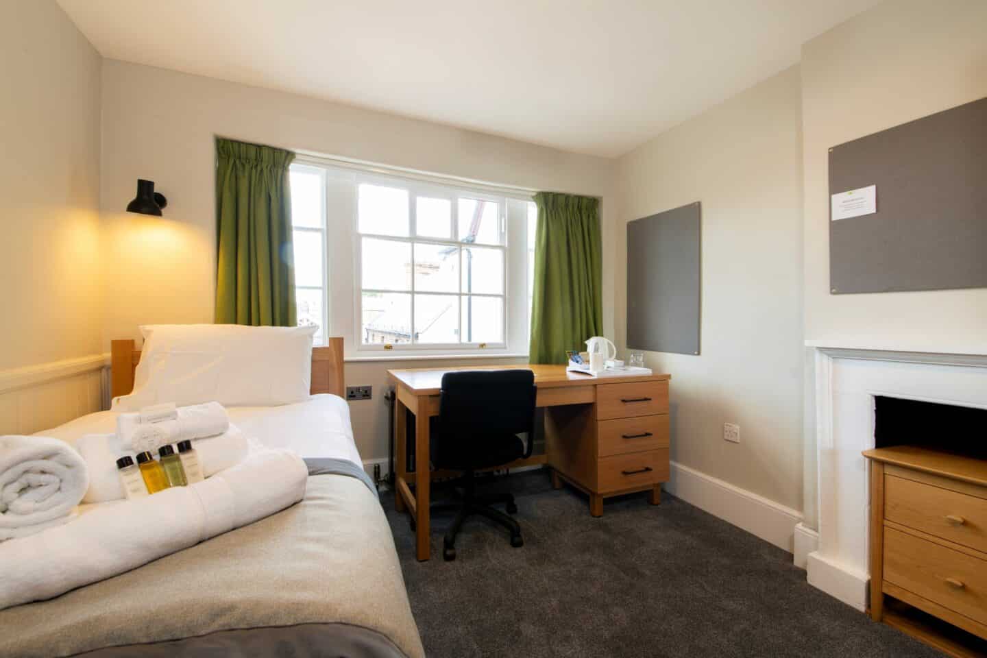 S06 - bedroom with a single bed (with pillow, sheets, towels, and toiletries), large windows with green curtains, a wooden desk with chair, drawers, and a kettle), a radiator, two corkboards, and a chest of drawers within a fireplace.