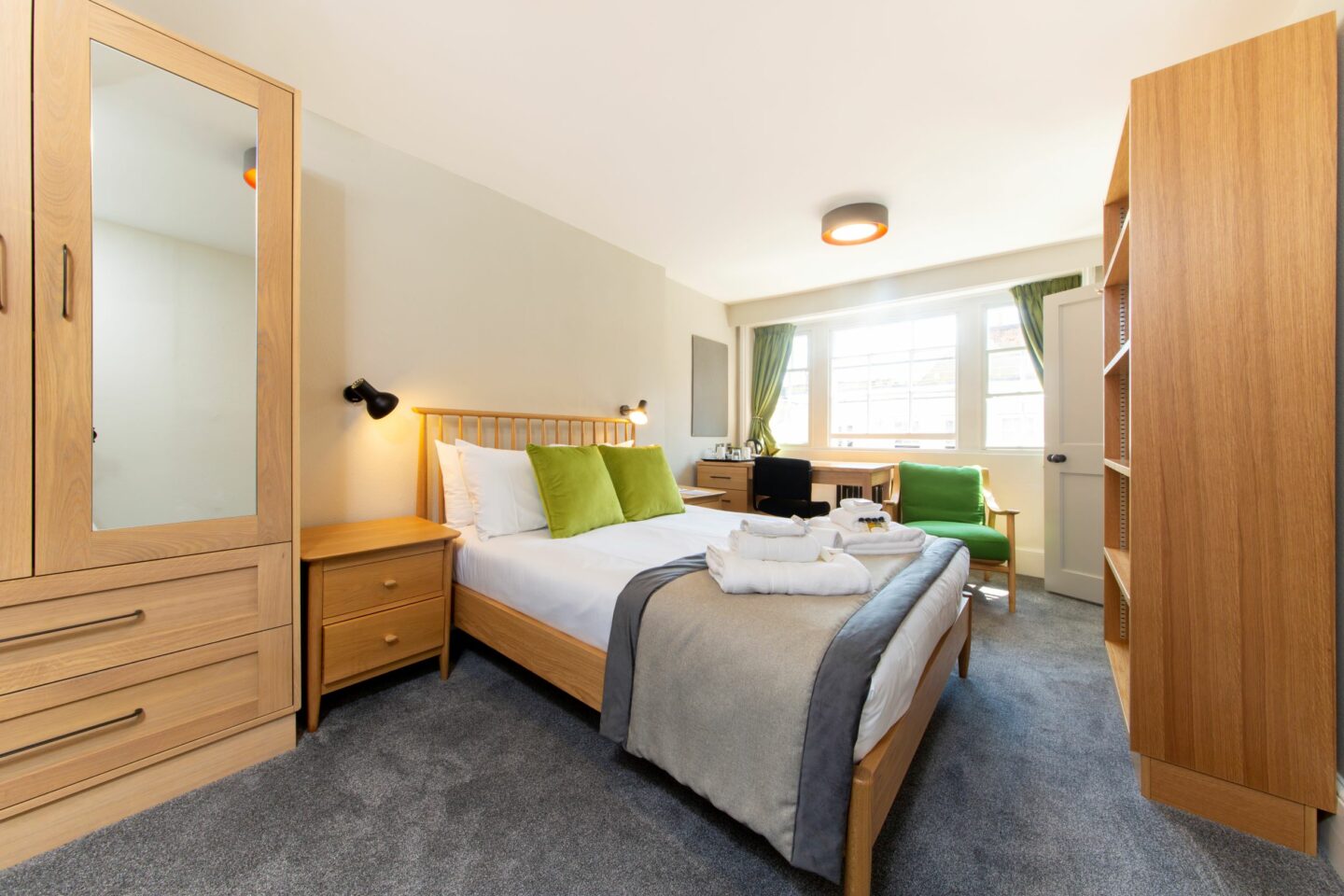 S01 - bedroom with a double bed (with sheets, pillows, cushions, towels, and toiletries), two small chests of drawers, a large wardrobe with a mirror, a large set of shelves, a wooden desk with chair, drawers and kettle, a corkboard, a radiator, an armchair, and large windows