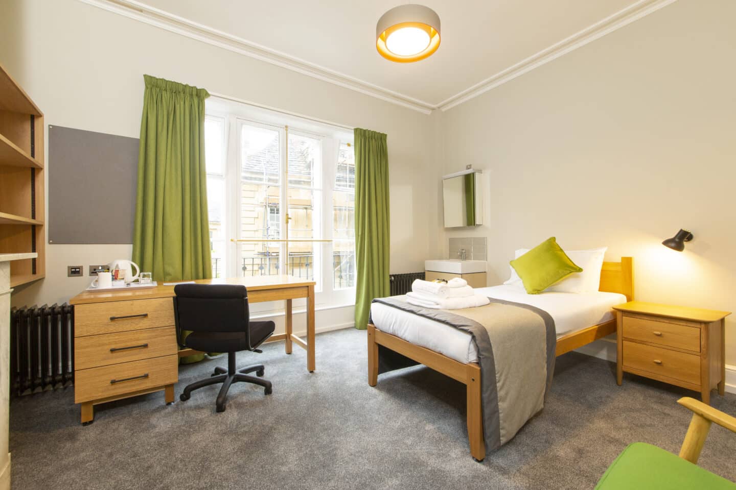 Bedroom with a single bed (with sheets, a pillow, a cushion, towels and toiletries), a sink and mirror, two radiators, a small chest of drawers, a wooden desk with drawers, chair, kettle and tea-set, a large set of windows with green curtains, a corkboard and shelves