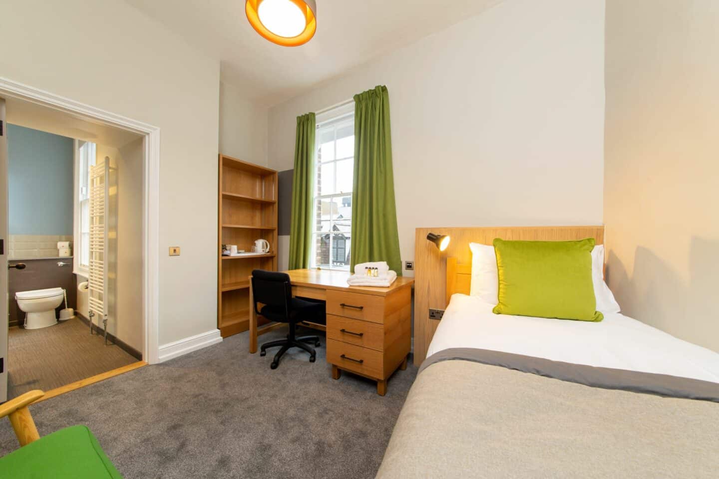 F75 - bedroom with single bed (with pillow, cushion, and sheets), a wooden desk with chair, drawers, towels and toiletries), a set of shelves with kettle and tea-set, a corkboard, a window with curtains, an armchair, and an en-suite bathroom with toilet and heated rack visible