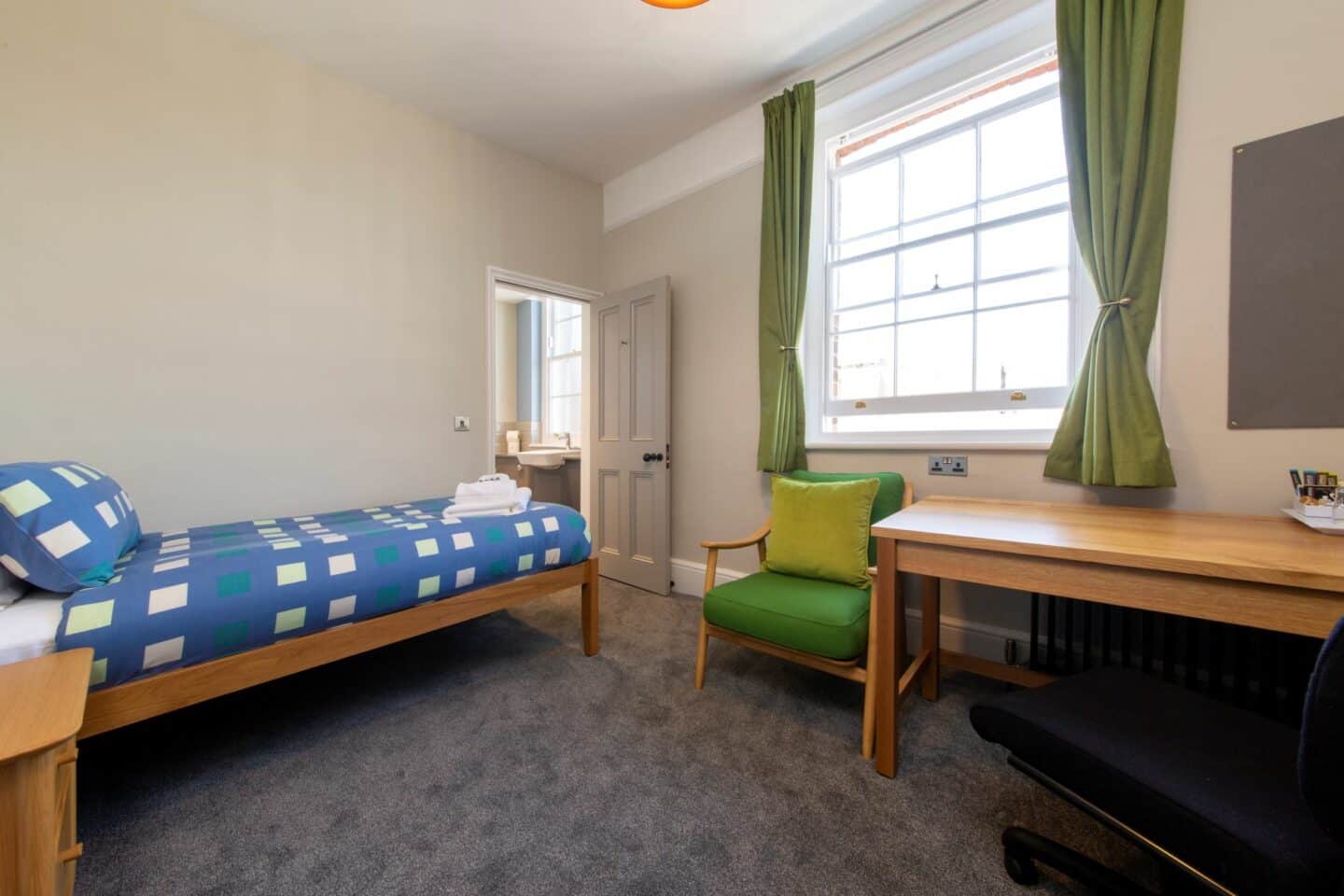 S86 - bedroom with a single bed (with blue sheets, a pillow, towels and toiletries), a wooden desk with chair, a radiator, an armchair and cushion, a corkboard, a large window with curtains, and an en-suite bathroom with a sink visible