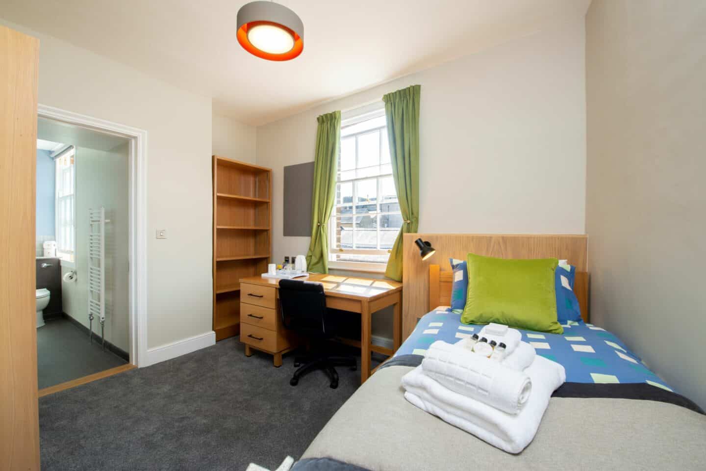 S94 - bedroom with a single bed (with pillows, a cushion, sheets, towels and toiletries, a wooden desk with chair, drawers, a kettle and tea-set, a window with curtains, a corkboard, a set of shelves, and an en-suite bathroom with toilet and heating rack visible