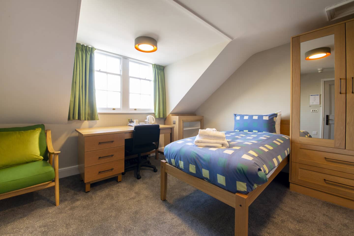 T44 - bedroom with a single bed (with towels and pillows), a wooden wardrobe with a mirror, small shelves, a wooden desk and chair (with a kettle and tea set), windows with green curtains, and a green armchair with a cushion