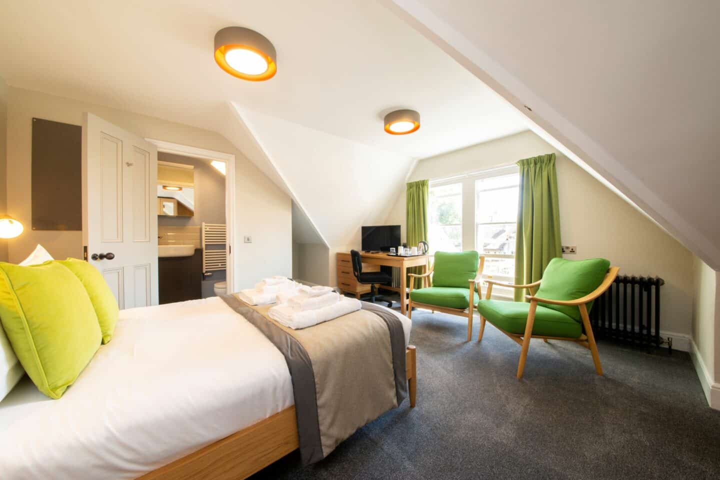 T43 - bedroom with a double bed (with sheets, pillows, cushions and towels), two green armchairs, a radiator, large windows with green curtains, a wooden desk with a chair, monitor (possibly a tv), and kettle, and an en-suite bathroom