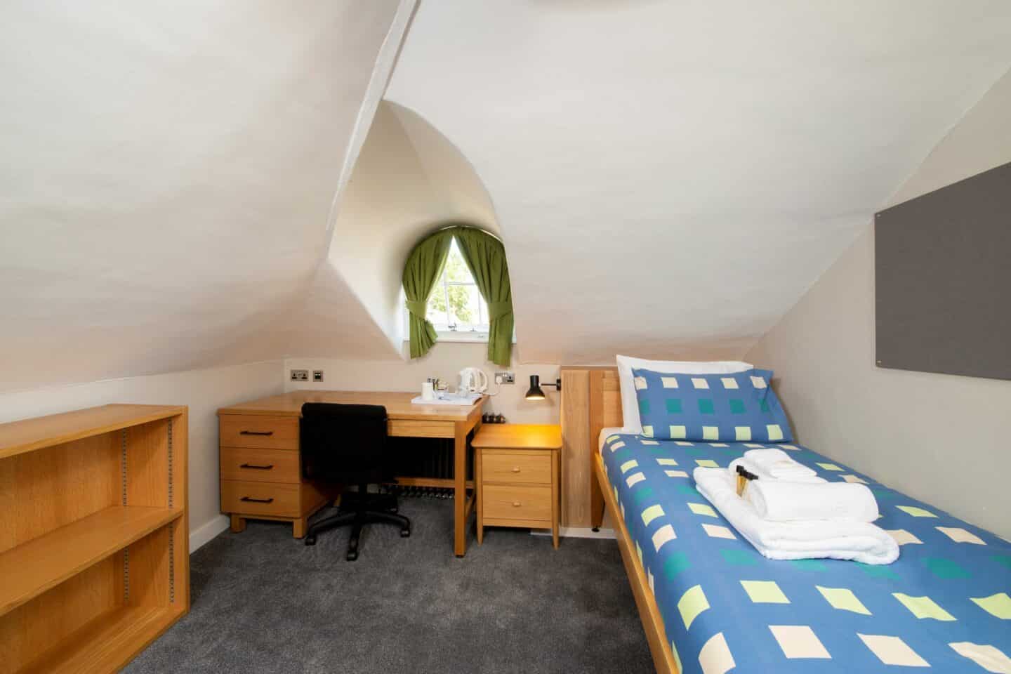 T47 - bedroom with a single bed (with pillows, sheets, and towels), a corkboard, shelves, a wooden desk with drawers, a chair, and a kettle, a smaller chest of drawers, a lamp, and a small window with green curtains