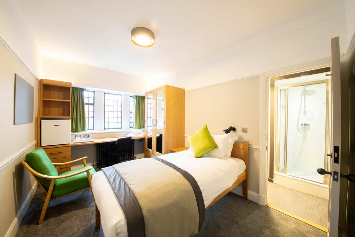 Bedroom with a single bed (with sheets, a pillow and cushion), a bedside table, an armchair, a wardrobe with mirrors and drawer, a chest of drawers and shelves, a corkboard, a desk with chair, a radiator, a kettle, towels, long windows with curtains, and an en-suite bathroom with a shower visible