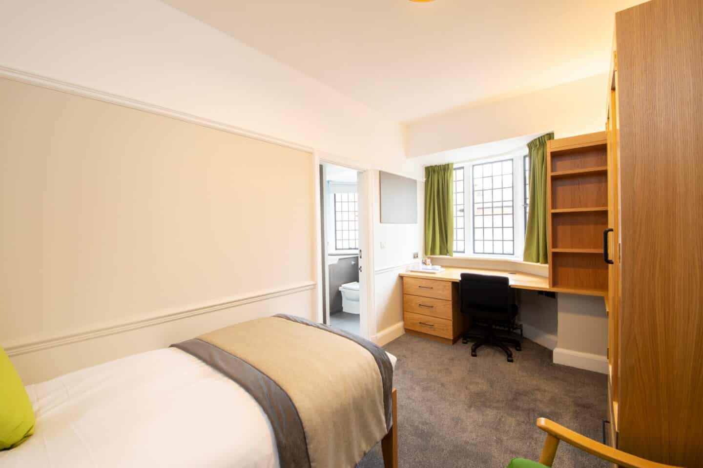 Bedroom with a single bed (with sheets and a cushion in frame), an armchair, a wardrobe, a wooden desk with chair, drawers, and a tea-set, a radiator, a corkboard, a set of shelves, large windows with curtains, and an en-suite bathroom with a toilet visible