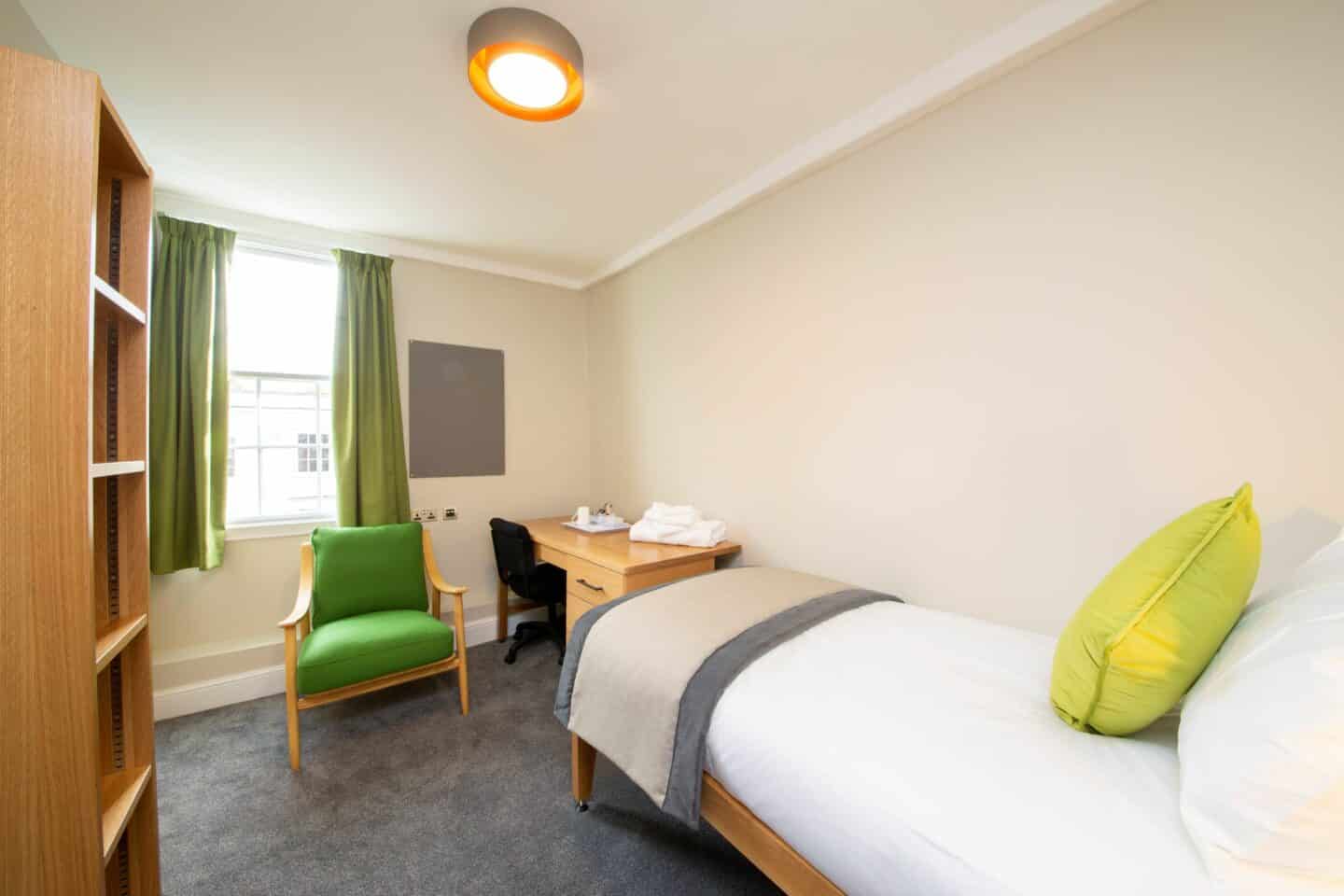 S14 - bedroom with a single bed (with sheets, a pillow and a cushion), a wooden desk with drawers, a chair, pillows, and a tea set, an armchair, a corkboard, a window with green curtains, and a large set of shelves