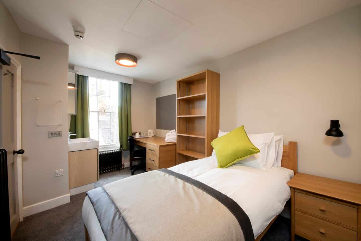 bedroom with a single bed (with sheets, pillows and a green cushion), a small chest of drawers, wooden shelves, a wooden desk and chair (with towels and a kettle and tea set), a corkboard, a window with green curtains, a radiator, and a sink