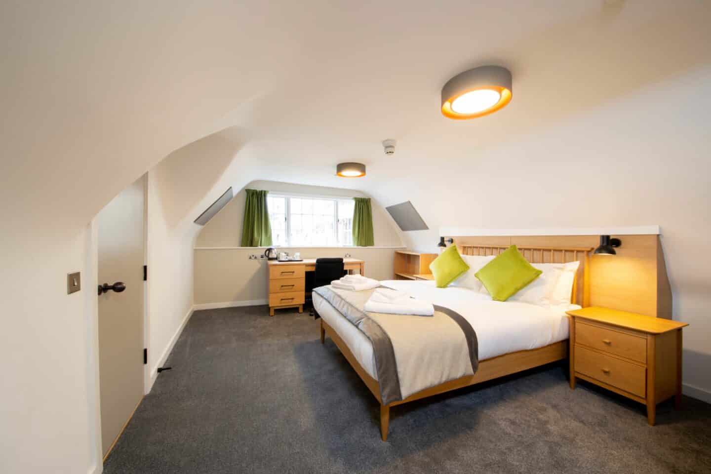Bedroom with a double bed (with sheets, towels, pillows, and green cushions), a small wooden chest of drawers, a wooden desk with chair and a kettle and tea set, a corkboard, and large windows with green curtains