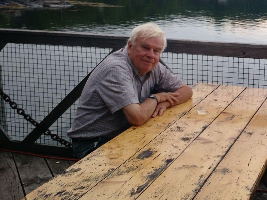 Photo of Stephen Dyson sitting at a rustic wooden table in Maine, USA. The ocean is sparkling behind him and he's leaning forwards with his arms resting on the table.