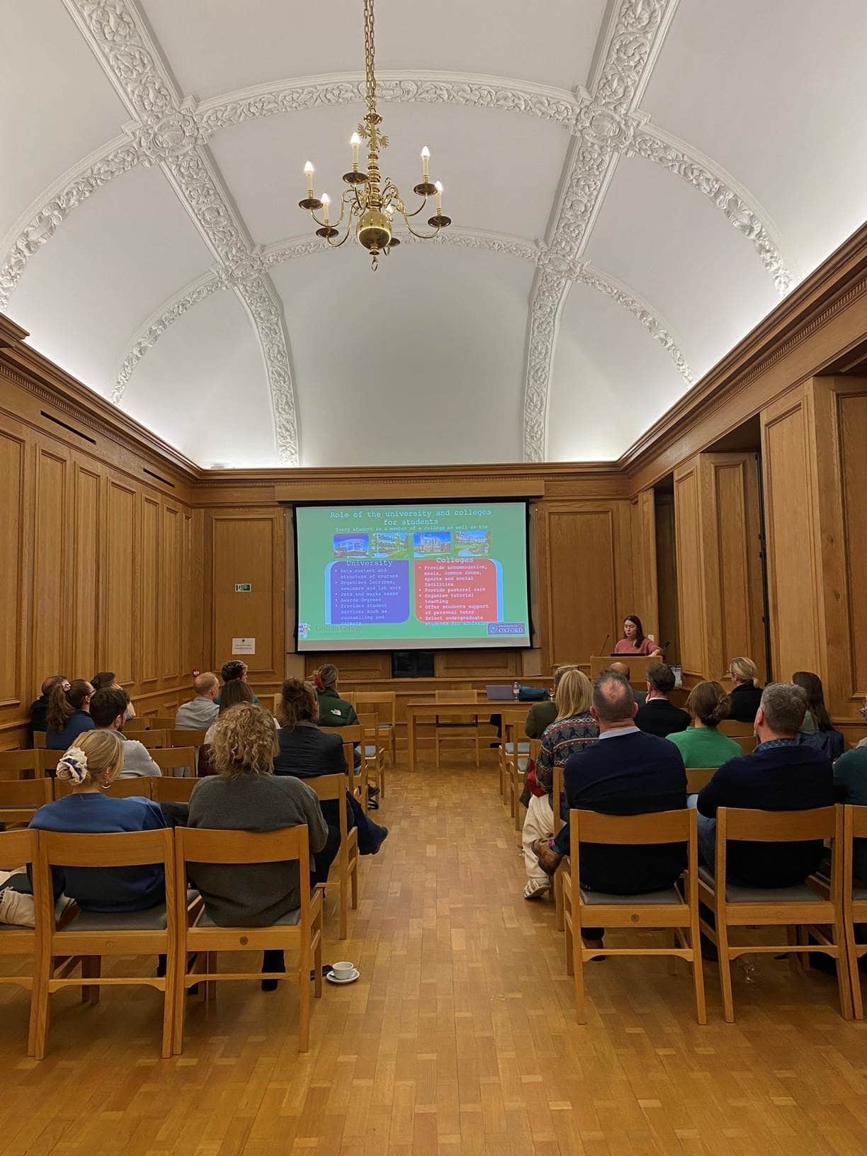 Participants sit in rows of chairs watching a presentation screen at the front of the room