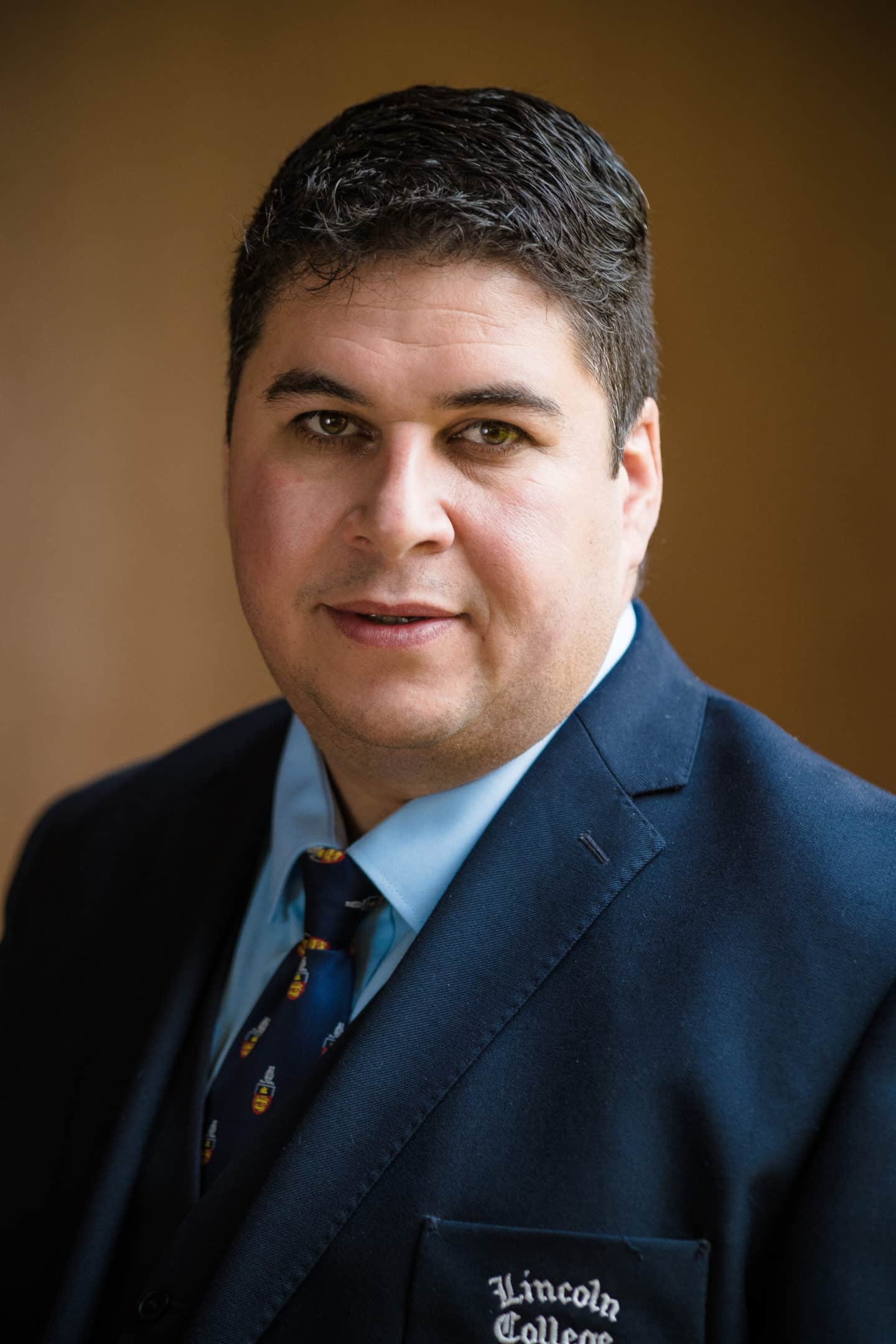 Headshot of Cristiano Da Silva, a man in a blue Lincoln College suit and tie