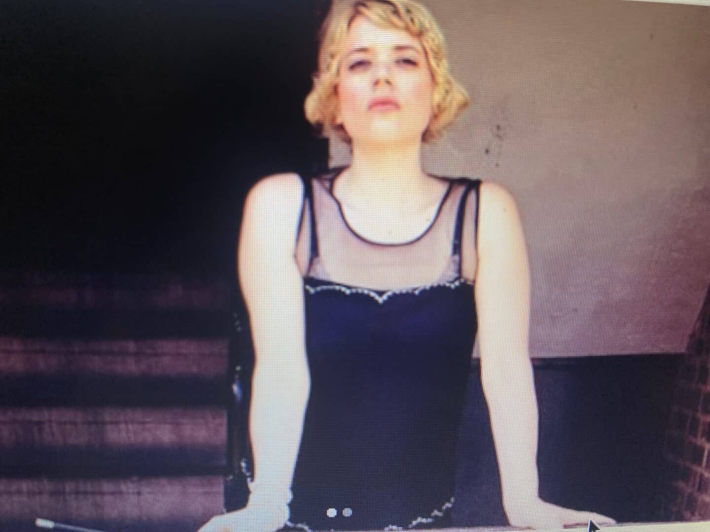 Beckis Cooper portraying Daisy Buchanan from The Great Gatsby. She stands confidently with her arms resting on a surface, wearing a 1920s-style black dress with sheer detailing and beaded embellishments. Her short, wavy blonde hair and bold expression capture the essence of the iconic character. The background is dimly lit, with stairs leading into the shadows, adding to the dramatic atmosphere.