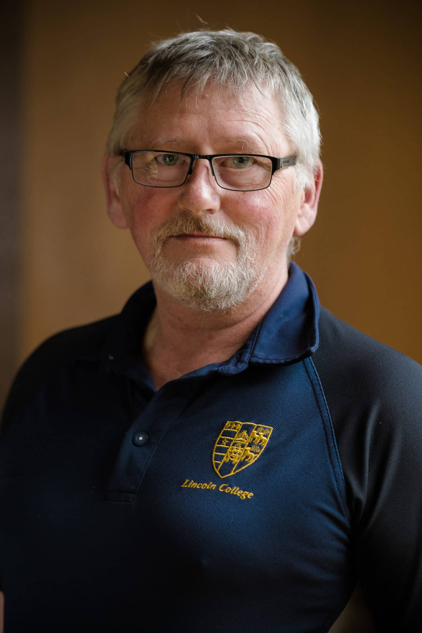 Headshot of David Harker, a man in glasses and a blue shirt with the Lincoln College crest in gold