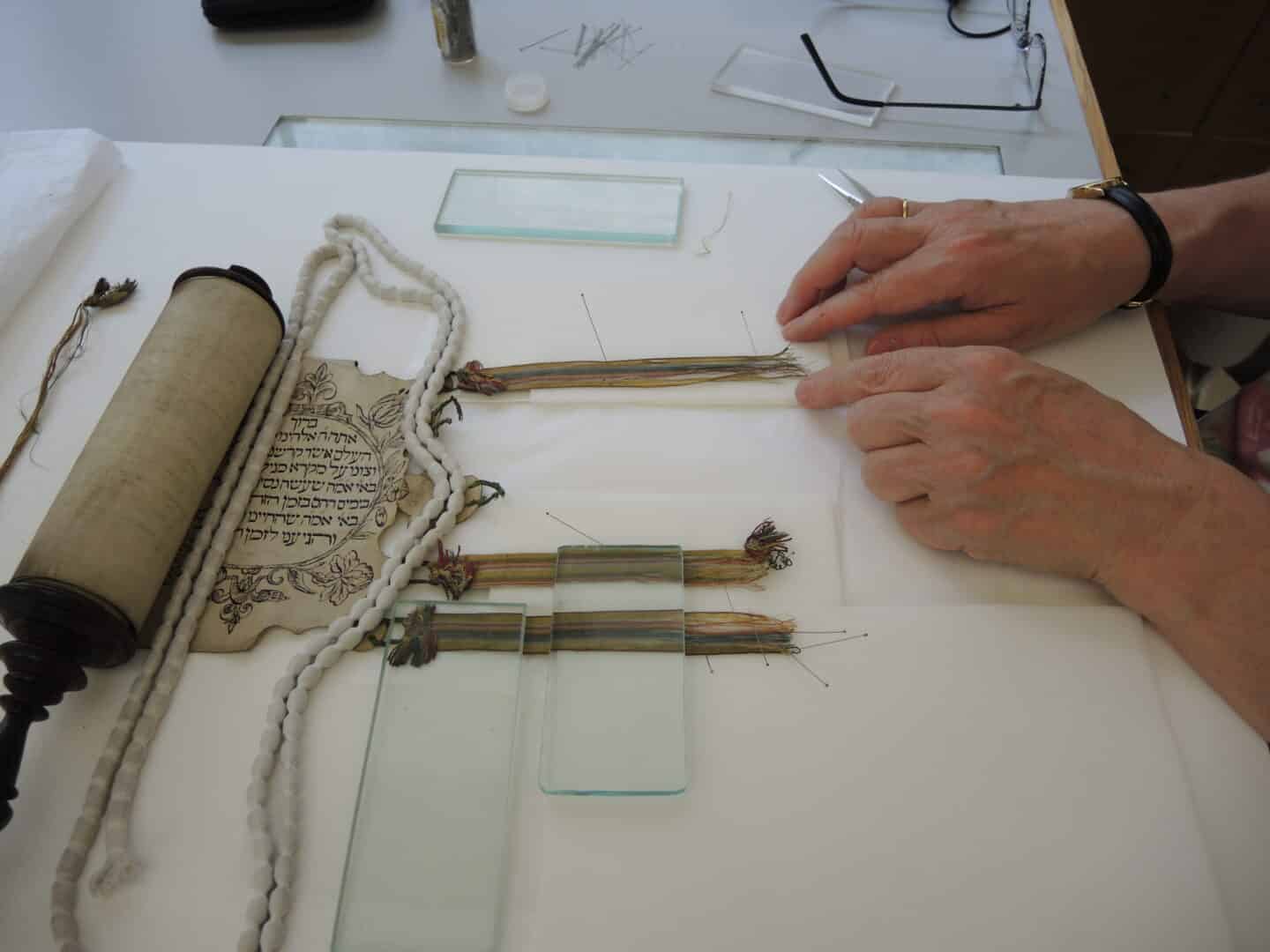 Partially unrolled roll with beautiful Hebrew script and three ornate woven ties of brightly coloured striped silk to tie the roll. The ties are covered in glass weights and have pins in them as they are being restored by a conservator whose hands are pictured