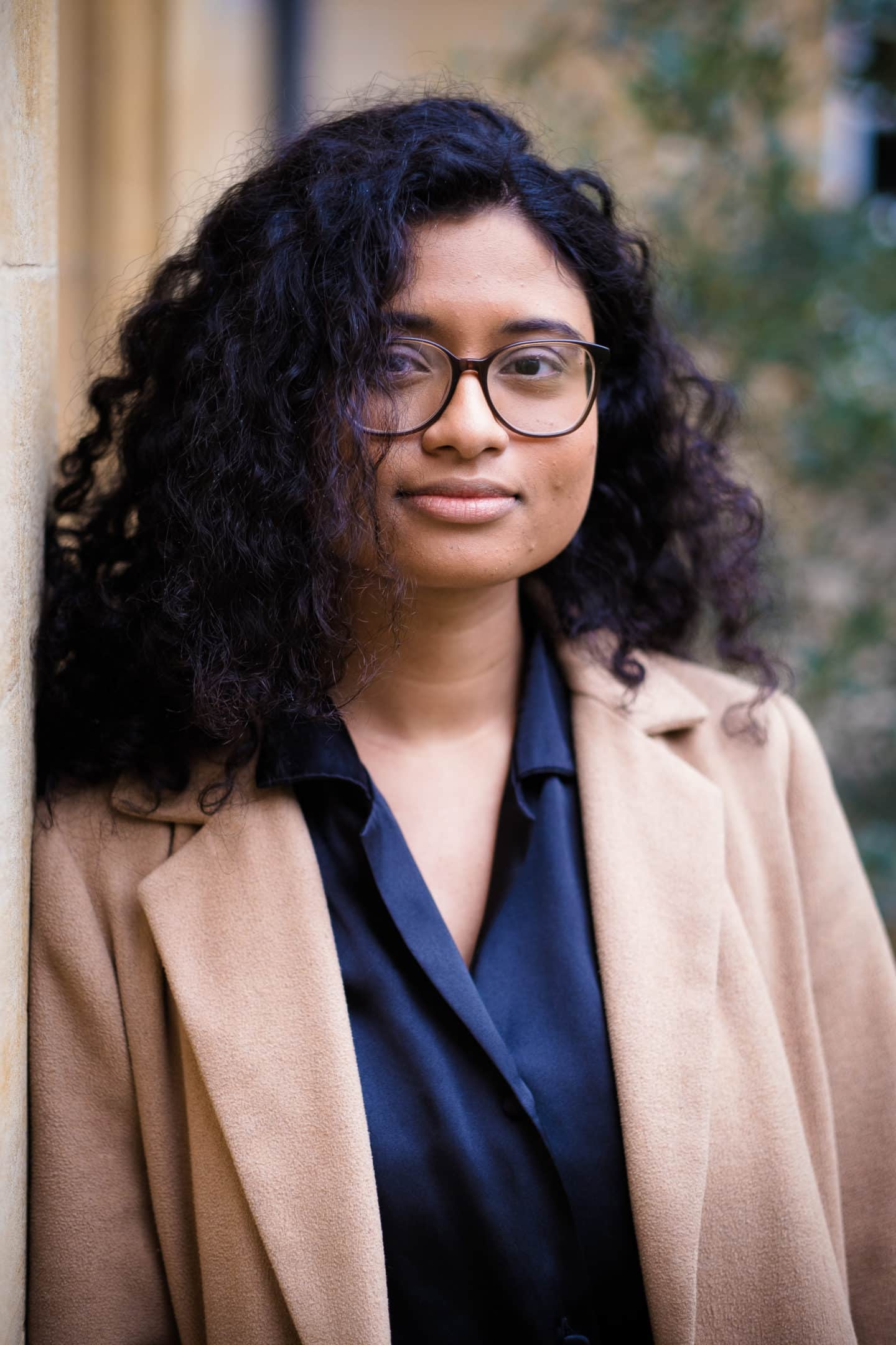 Headshot of current student, Gayatri Tadikamalla, a young woman in a blue top, brown coat, and glasses