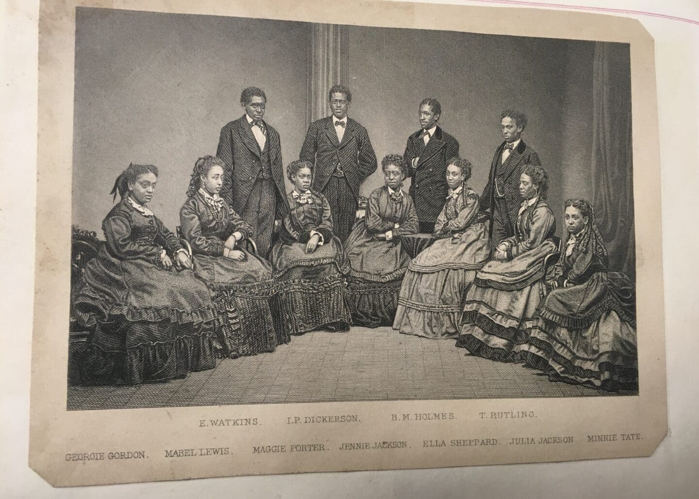 Black and white image of a group of people, seven women in long dresses sitting on chairs with four men in suits and bowties standing around them