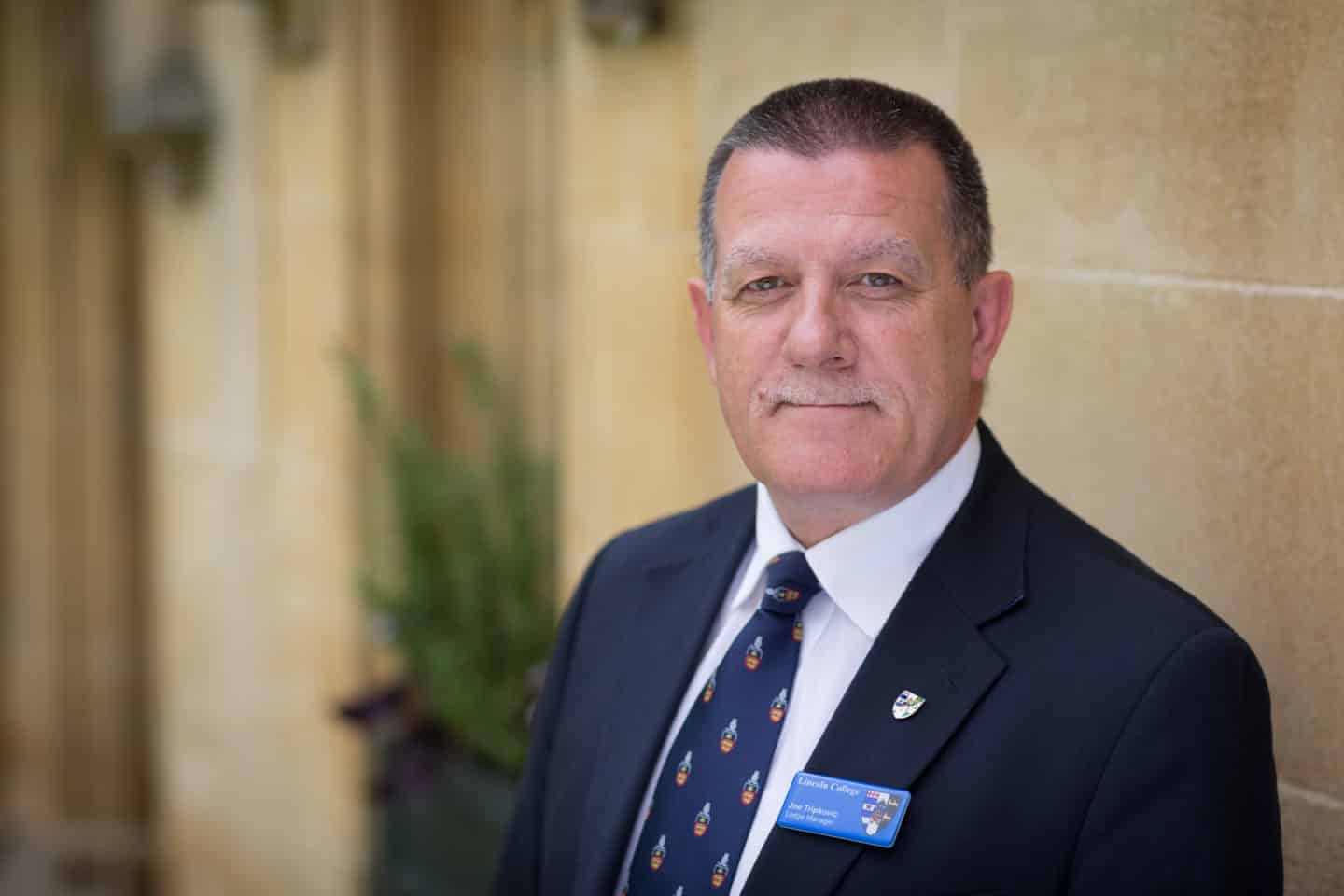 Headshot of the Lodge Manager Joe Tripkovic, a man in a suit and tie with a Lincoln College badge
