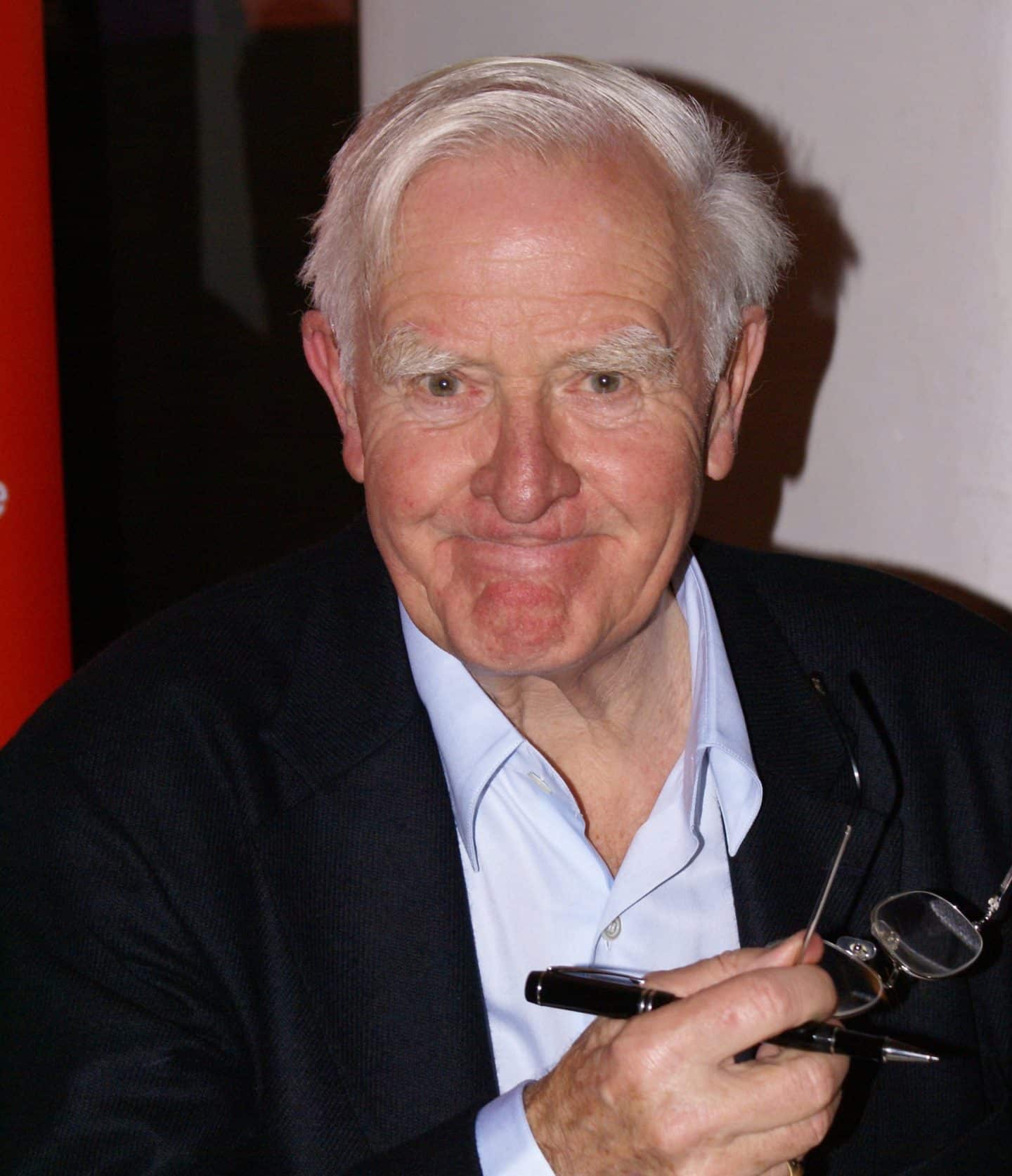 Photograph of John le Carre, a man wearing a suit and holding his glasses and a pen in one hand