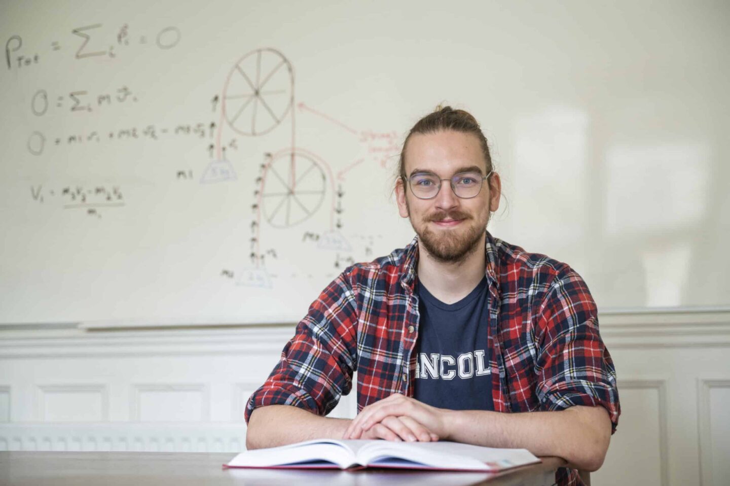 Photograph of Joshua Green seated at a table with a whiteboard with equations in the background