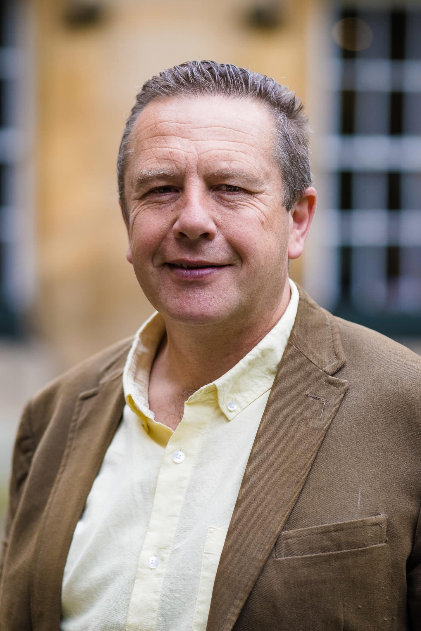 Headshot of Julian Mitchell, a man in a pale yellow shirt and brown jacket