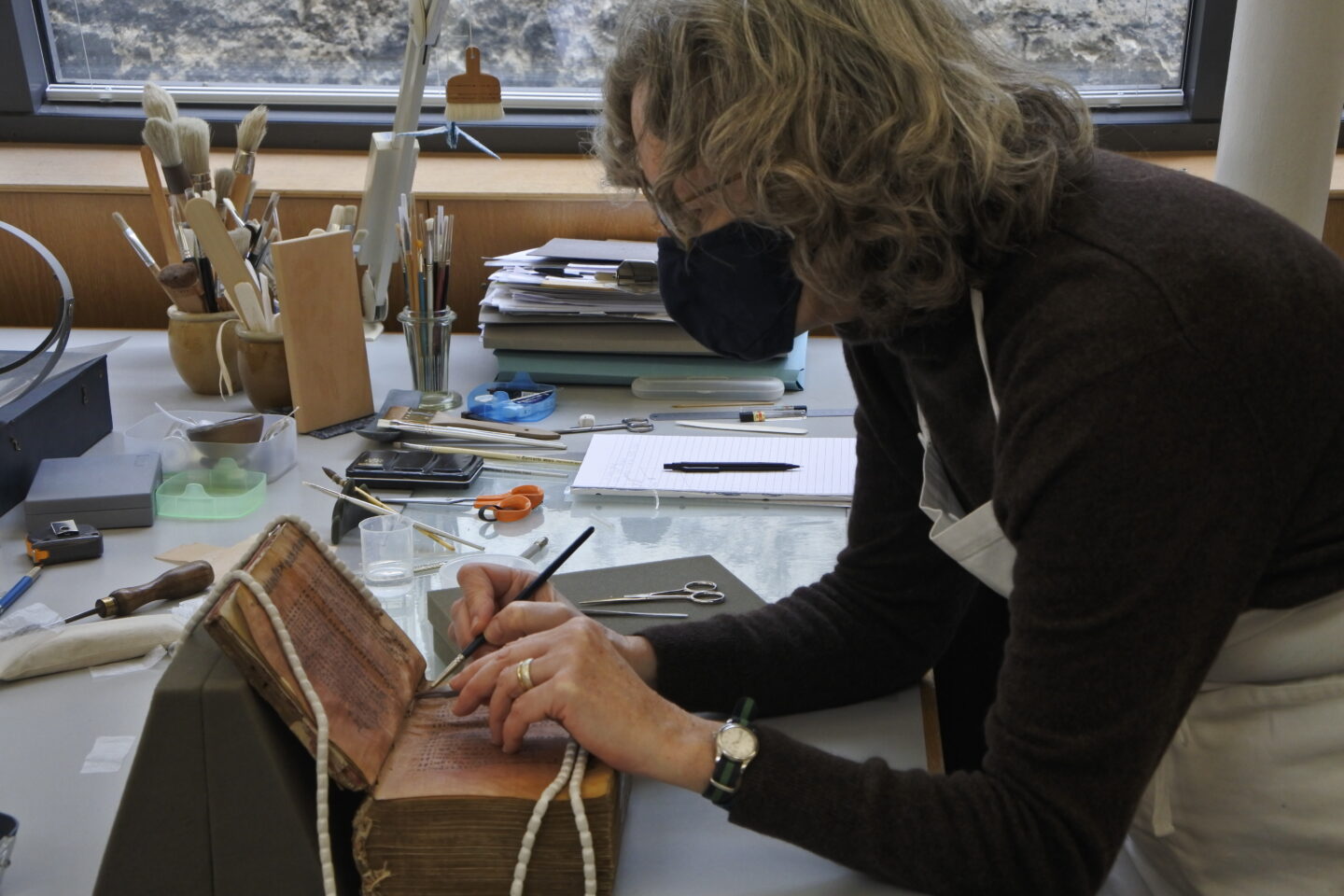 A woman in a black mask is bent over an old open manuscript, restoring it with tools (including a paintbrush). The table is covered in a variety of other tools.