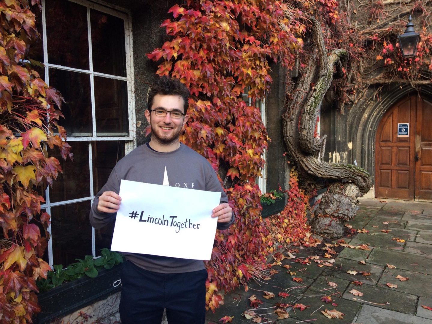 A young man standing in Lincoln College's Front Quad with red ivy in the background, holding up a sheet of paper that reads #LincolnTogether