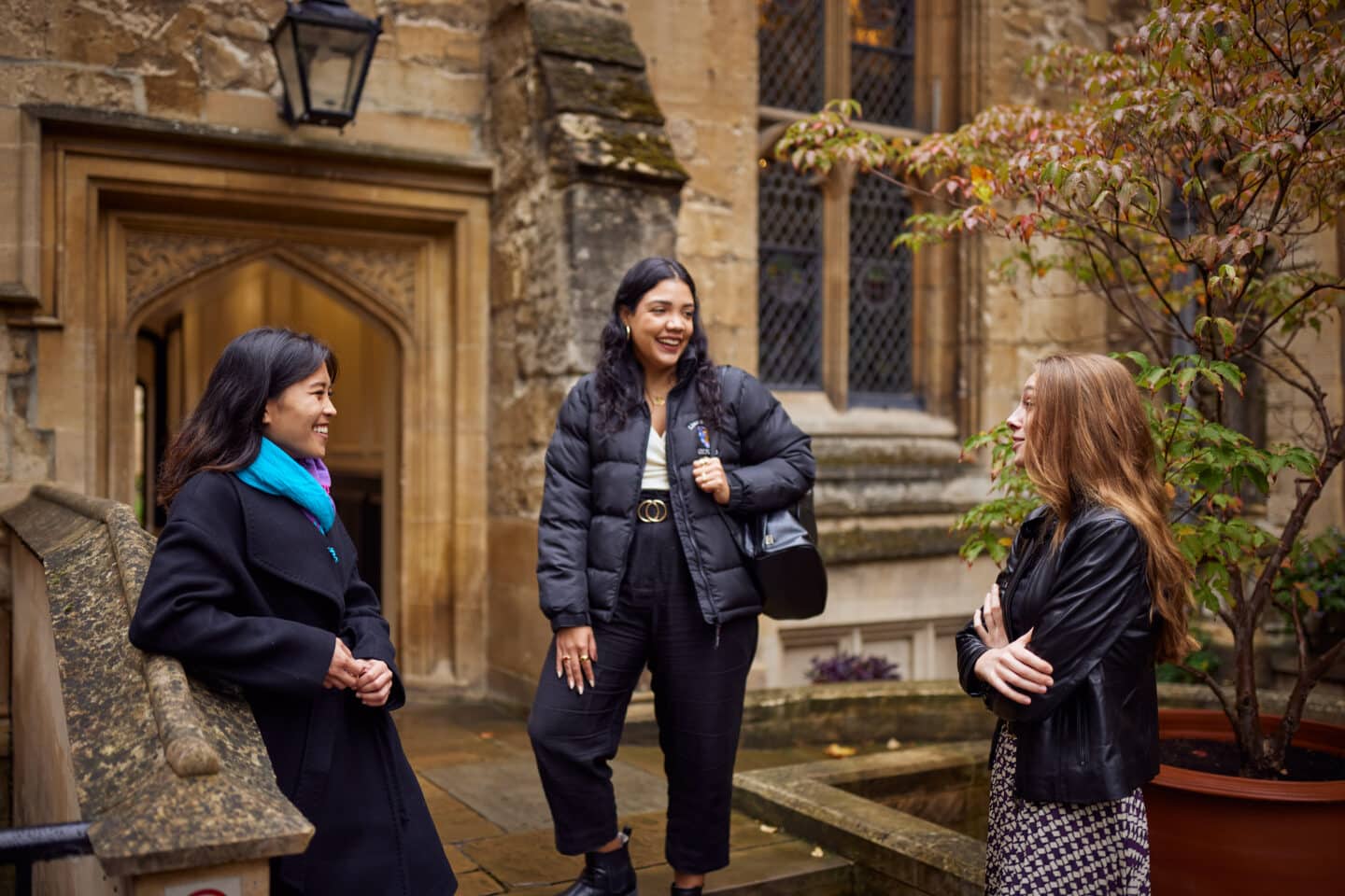 Three female students standing on the steps by an old stone arched doorway in Lincoln College, smiling and chatting